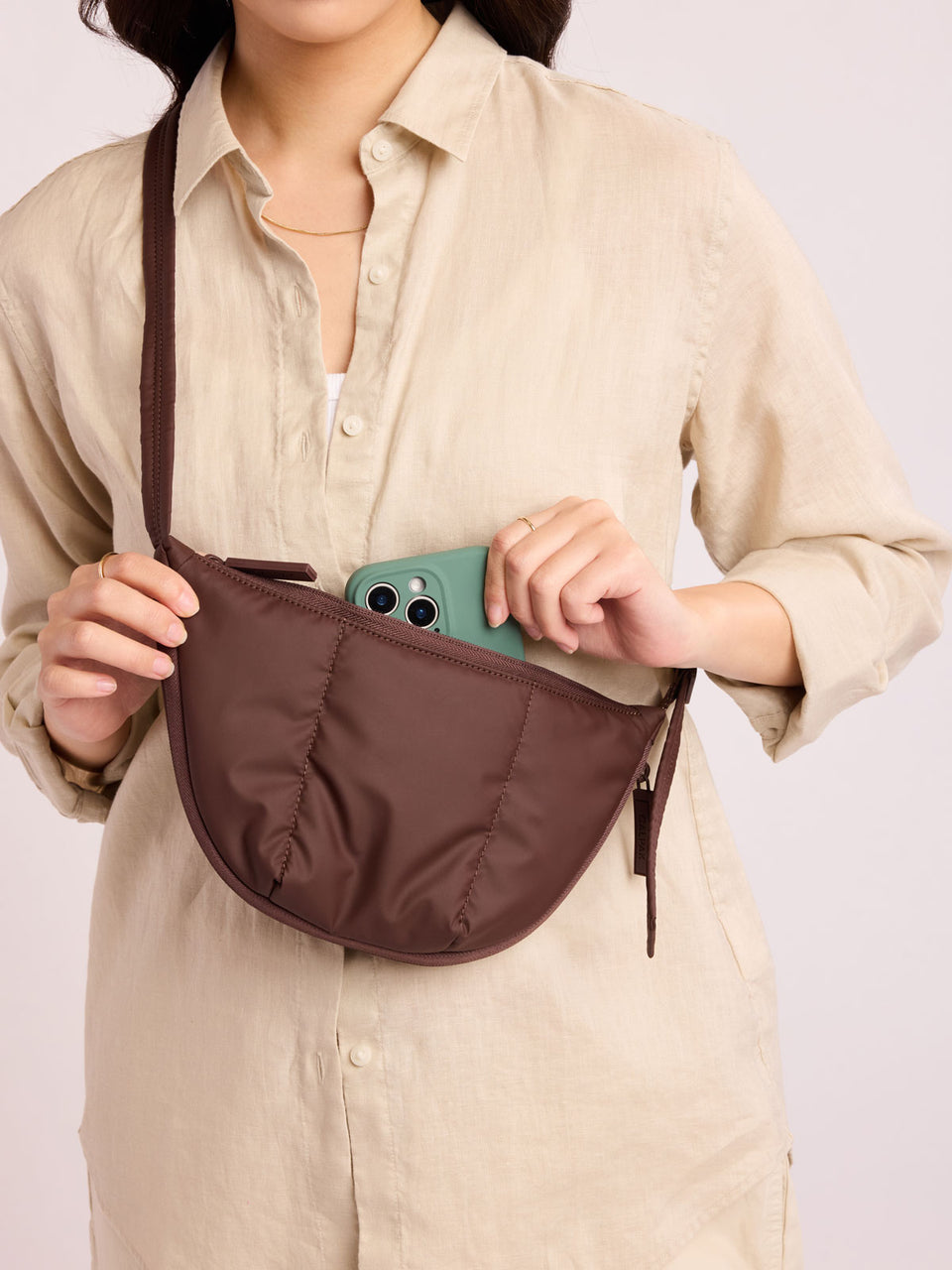 Person with beige outfit putting green phone into CALPAK Luka Crescent Tote bag in brown Walnut with shoulder strap and bag is expandable up to 2" on a white background; CBL2601-WALNUT