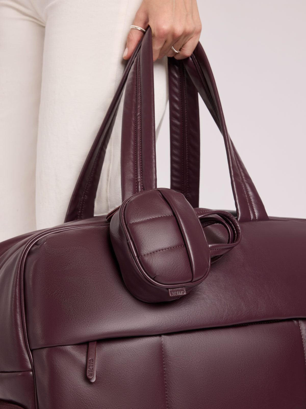 A close-up of a person in white pants holding a CALPAK Luka Duffel in Dark Cherry Vegan Leather with a matching small CALPAK Luka Duffel Zip Bag Charm in Dark Cherry Vegan Leather attached; ALK2501-DARK-CHERRY-PU