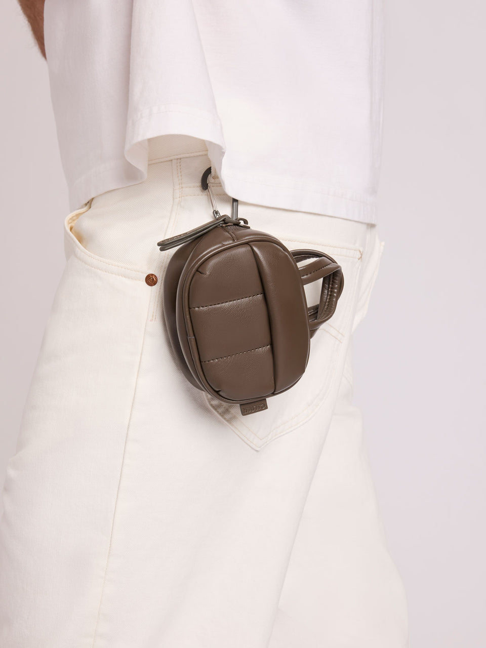 A dark brown Mushroom vegan leather top-handle CALPAK Luka Zip Bag Charm with a zippered front pocket and a small side strap, worn on white pants; ALK2501-MUSHROOM-PU