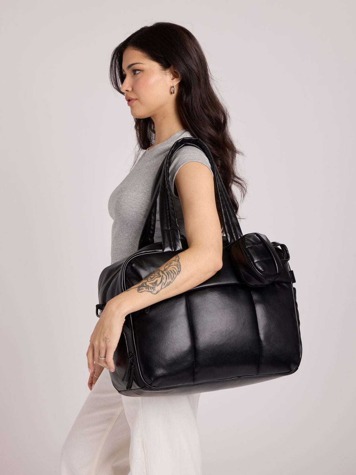 A woman in a gray sleeveless top and white pants carries a Black CALPAK Luka Duffel in vegan leather with dual top handles and a front zippered pocket, photographed against a clean white background; DSM1901-BLACK-PU