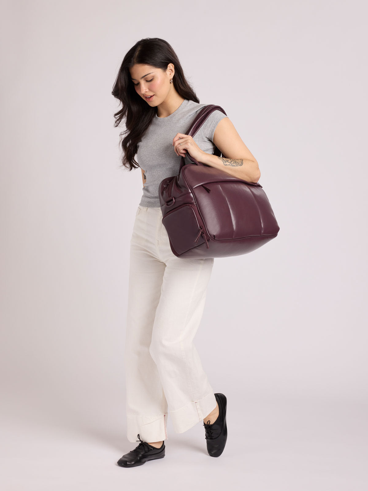 A woman wearing a gray sleeveless top, white pants, and black shoes carries a  dark burgundy CALPAK Luka Duffel in vegan leather with dual top handles and a front zippered pocket, photographed against a clean white background; DSM1901-DARK-CHERRY-PU