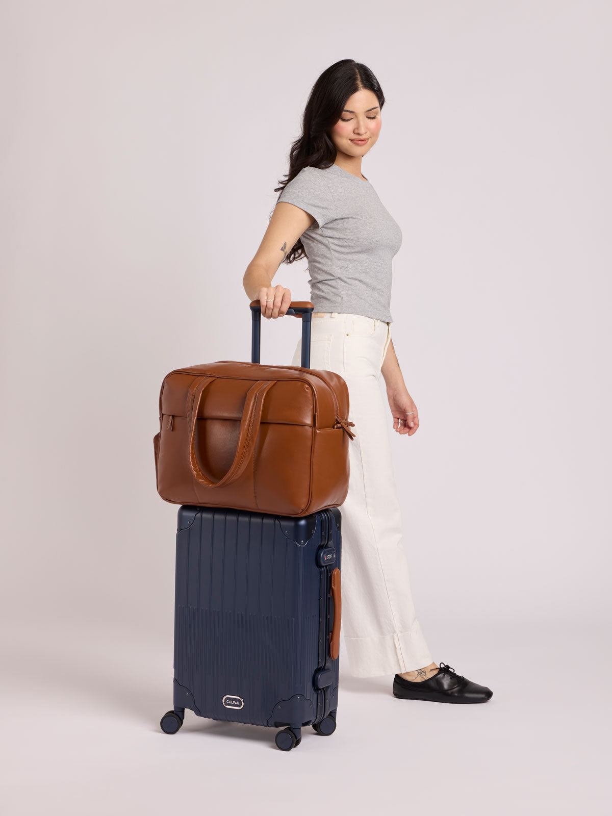 A woman stands in a studio holding the handle of a CALPAK Vault Luggage in Navy blue rolling suitcase, with a Honey Brown CALPAK Luka Duffel in vegan leather with dual top handles and a front zippered pocket, photographed against a clean white background; DSM1901-HONEY-BROWN-PU