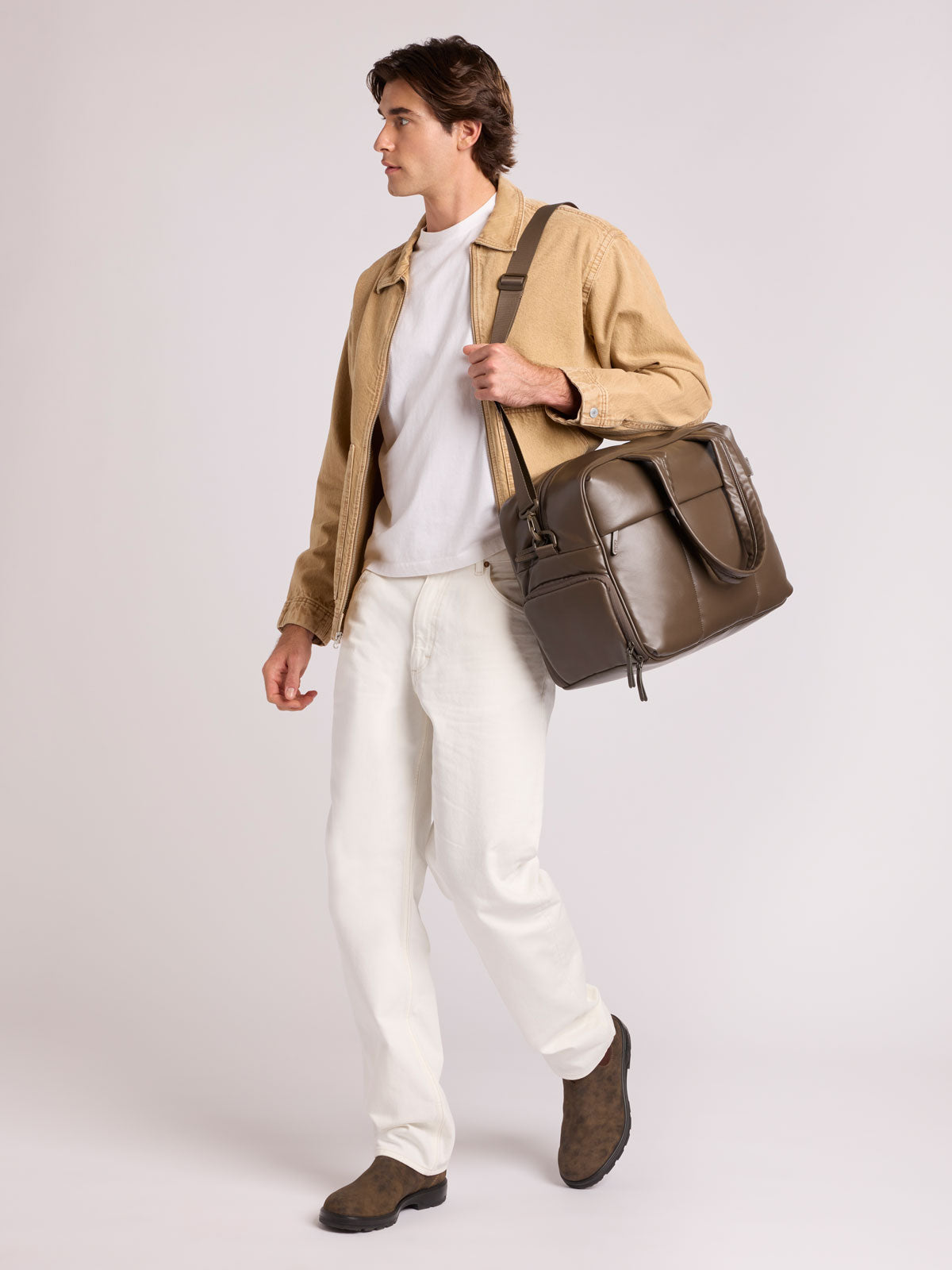 A man wearing a tan jacket, white t-shirt, white trousers, and brown boots carries a  dark brown Mushroom CALPAK Luka Duffel in vegan leather with dual top handles and a front zippered pocket, photographed against a clean white background; DSM1901-MUSHROOM-PU