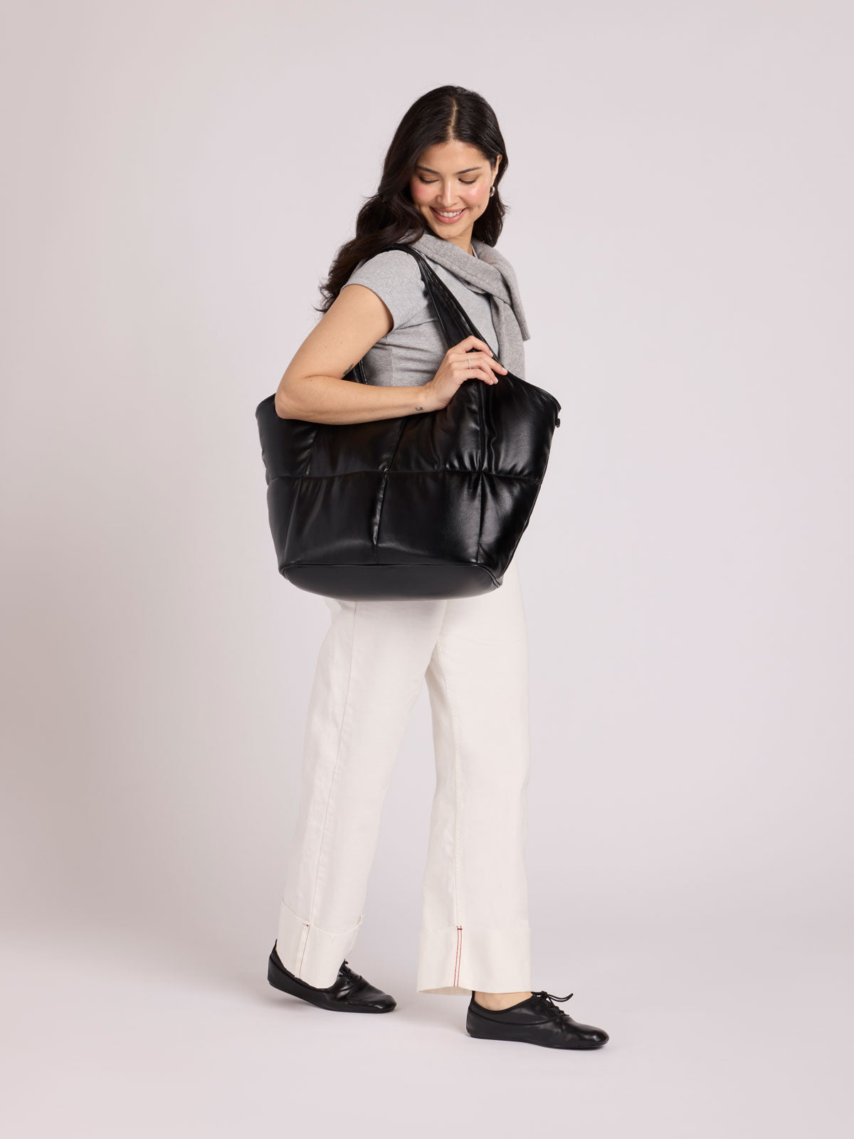 A woman modeling a CALPAK Luka Everything Tote in Black Vegan Leather, wearing a gray sleeveless top, white pants, and black shoes against a light gray studio background; AET2601-BLACK-PU