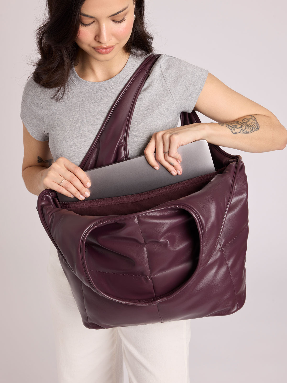 A woman wearing a gray t-shirt and white pants wears a CALPAK Luka Everything Tote Bag in Dark Cherry vegan leather, demonstrating its spacious interior with a gray laptop; AET2601-DARK-CHERRY-PU