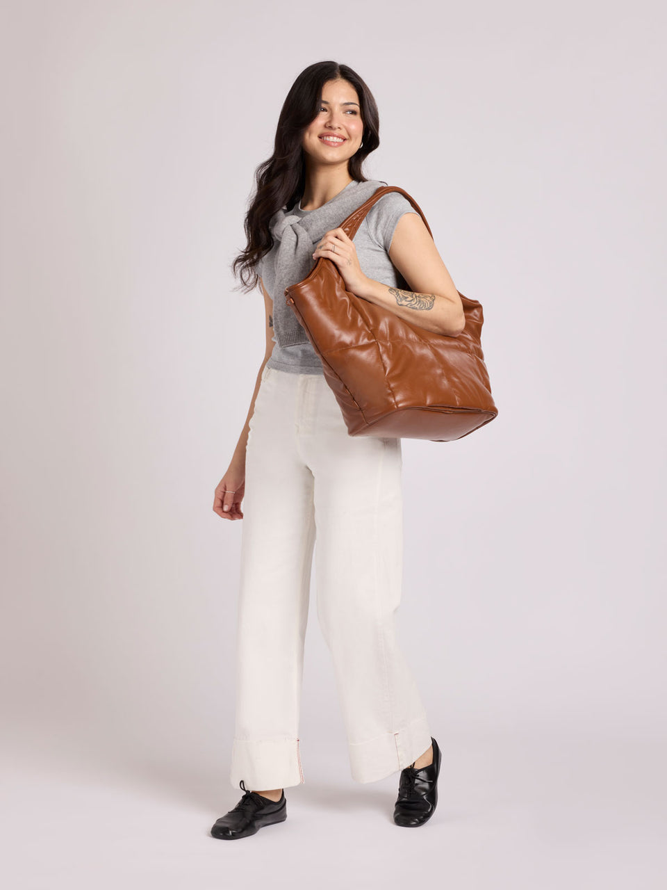 A woman modeling a CALPAK Luka Everything Tote in Honey Brown Vegan Leather over her shoulder, wearing a gray sleeveless top, white wide-leg pants, and black loafers, against a light gray studio background; AET2601-HONEY-BROWN-PU