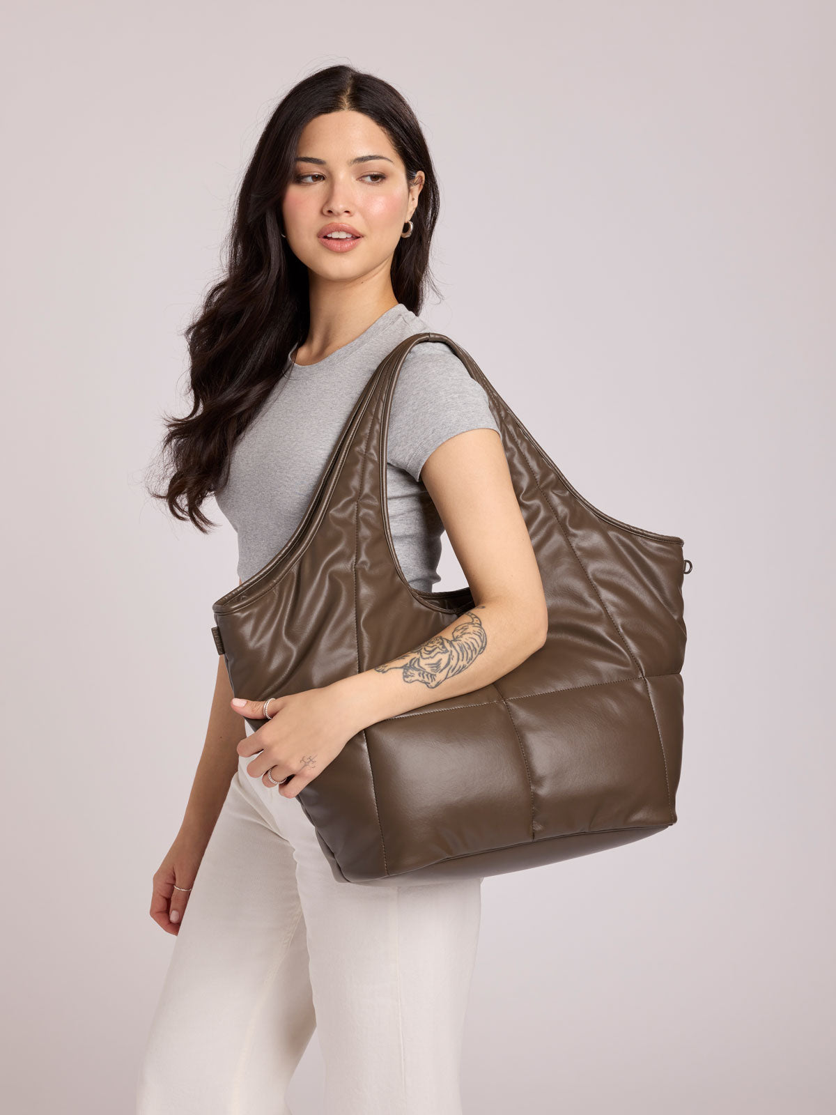 A woman wearing a gray t-shirt and white pants carries a CALPAK Luka Everything Tote in Mushroom brown Vegan Leather over her shoulder against a light gray studio background; AET2601-MUSHROOM-PU