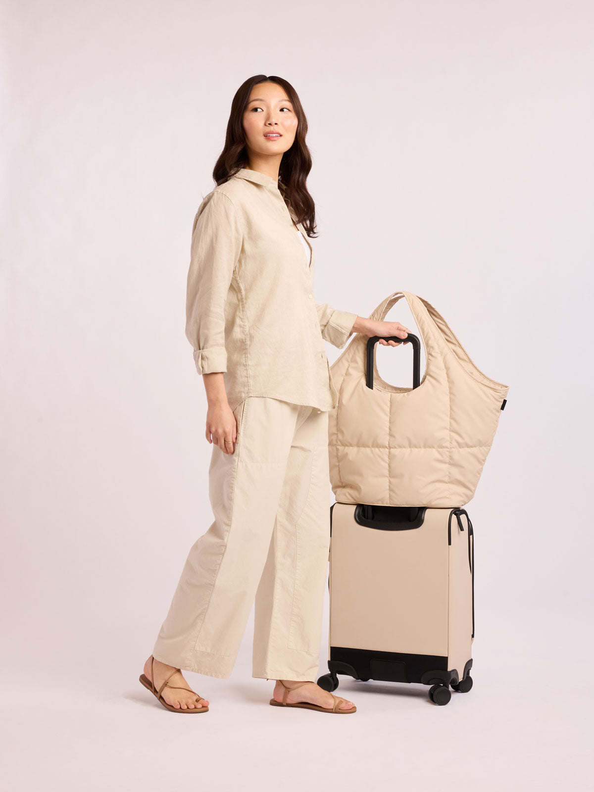 Model with CALPAK Luka Everything Tote in off-white beige Oatmeal on top of a suitcase with trolley luggage sleeve; AET2601-OATMEAL