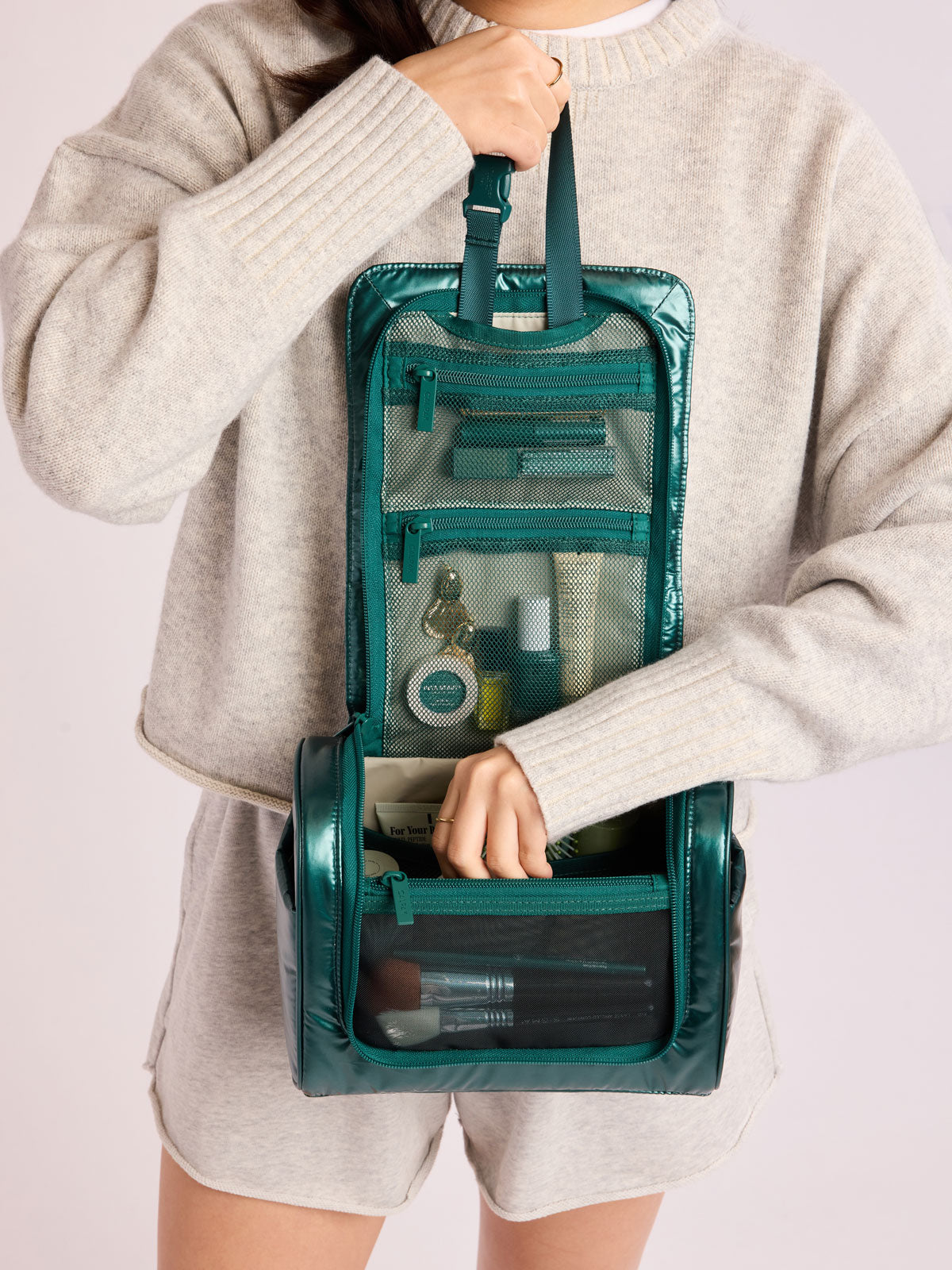 Person adding toiletries in CALPAK Luka Hanging Toiletry Bag in dark green shiny Kale; TLH2301-GLAZED-KALE