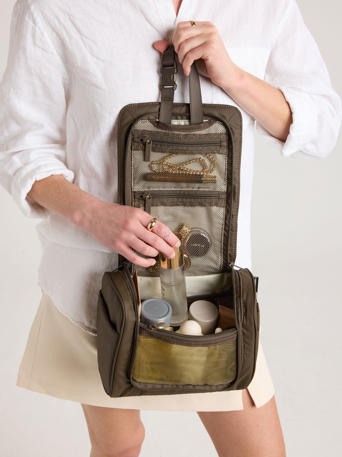 Person adding toiletries into CALPAK Luka Hanging Toiletry Bag in brown Mushroom; TLH2301-MUSHROOM