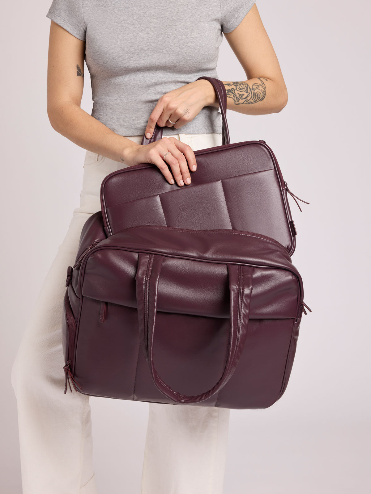 Woman wearing a grey top and white pants putting a CALPAK Luka 14in Padded Laptop Sleeve in Dark Cherry Vegan Leather into a CALPAK Luka Duffel in Dark Cherry Vegan leather against a plain background; ALL2401-DARK-CHERRY-PU
