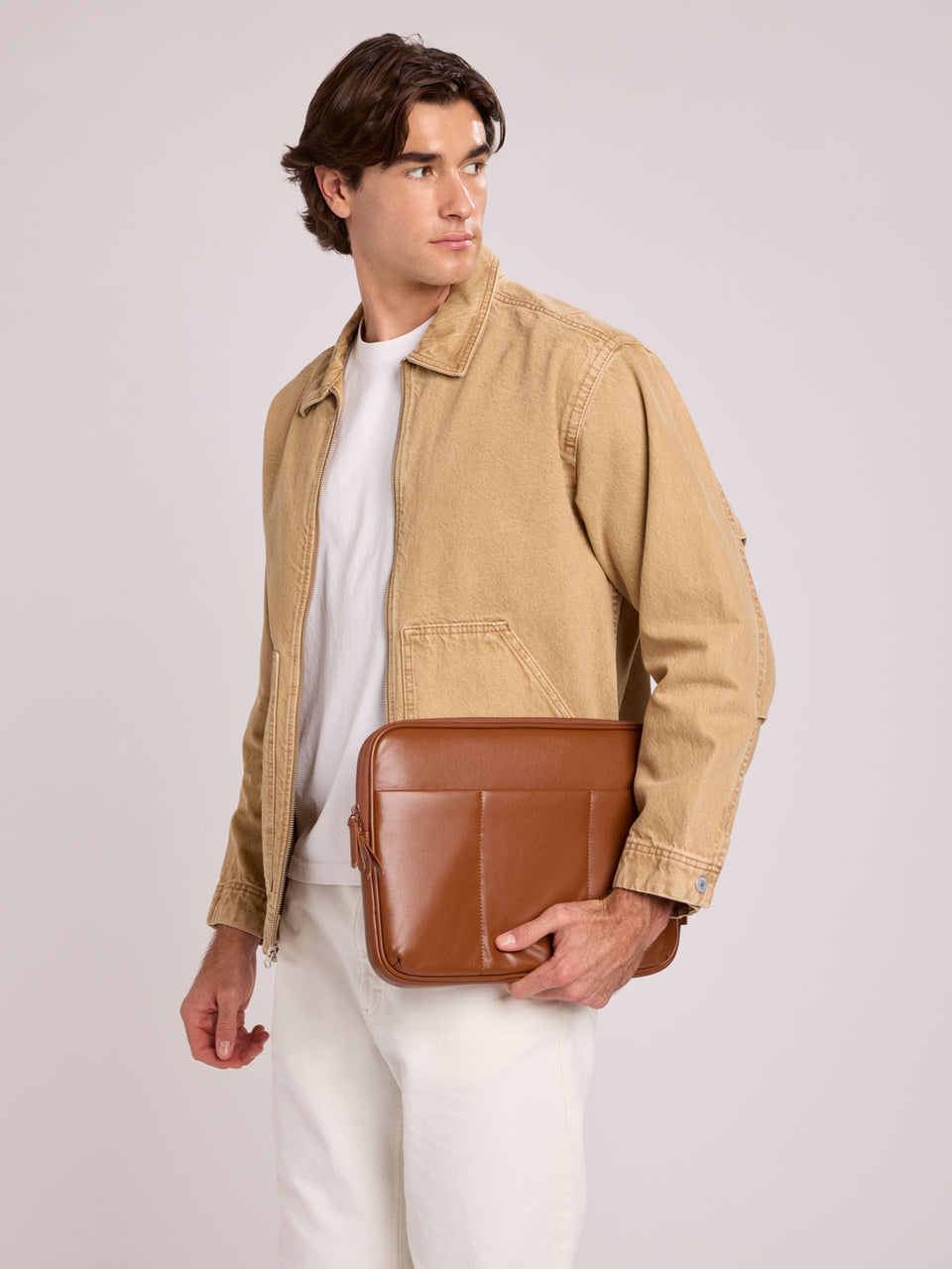 A man wearing a tan canvas utility jacket over a white t-shirt and white pants, holding a CALPAK Luka 14in Padded Laptop Sleeve in Honey Brown Vegan Leather; ALL2401-HONEY-BROWN-PU