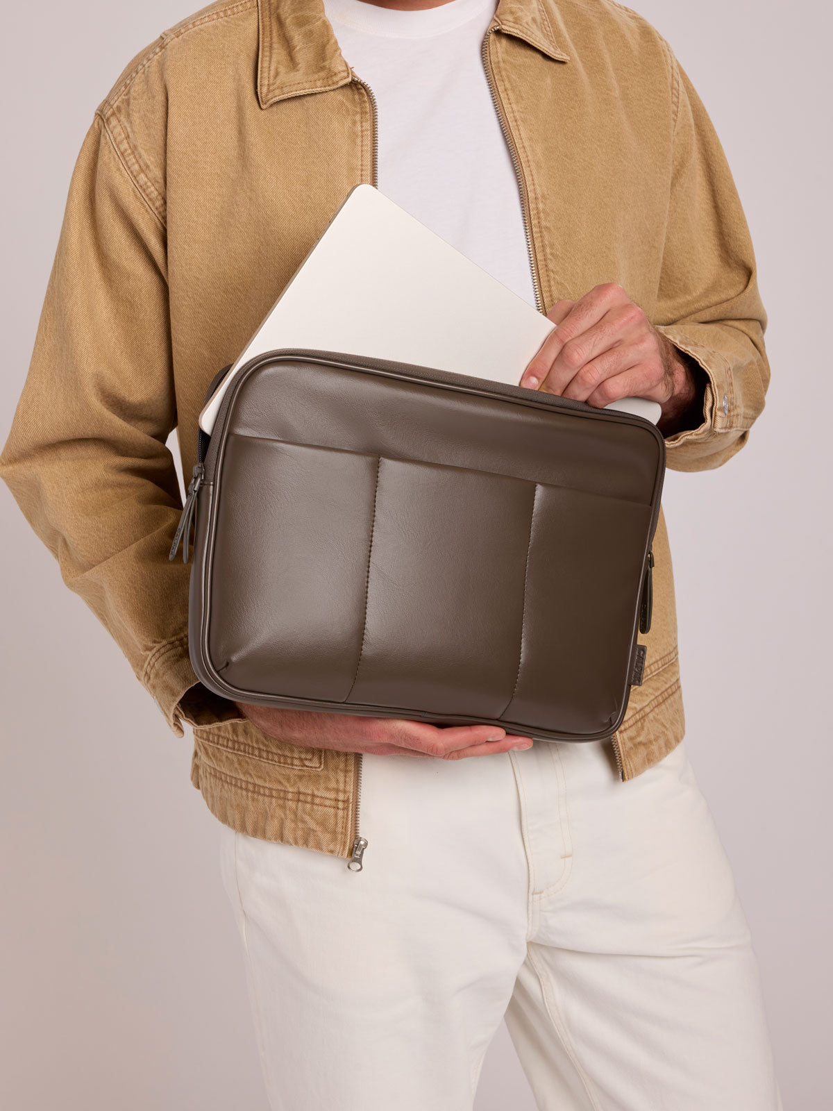 A person wearing a tan jacket and white pants holds a CALPAK Luka 14in Laptop Sleeve in Mushroom brown Vegan Leather with a white laptop inside, against a light gray studio background; ALL2401-MUSHROOM-PU