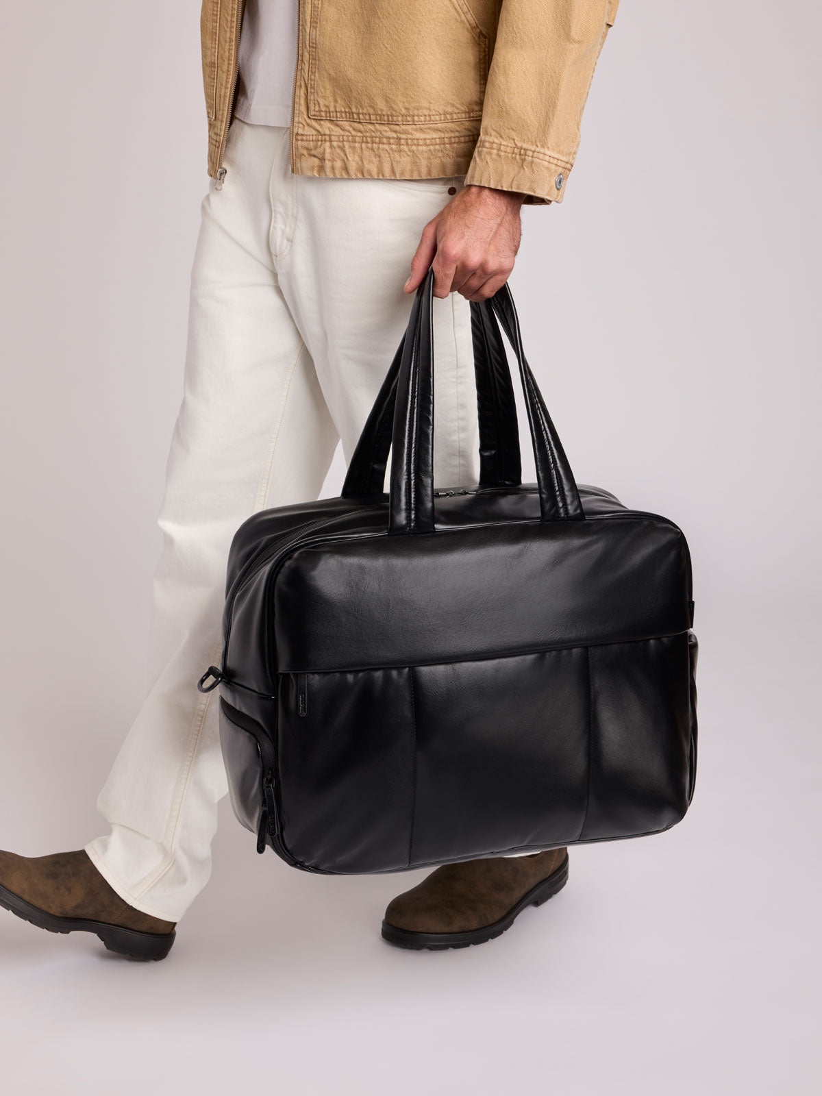 A person in a tan jacket, white pants, and brown shoes carries a Black CALPAK Luka Duffel in vegan leather with dual top handles and a front zippered pocket, photographed against a clean white background; DLL2201-BLACK-PU
