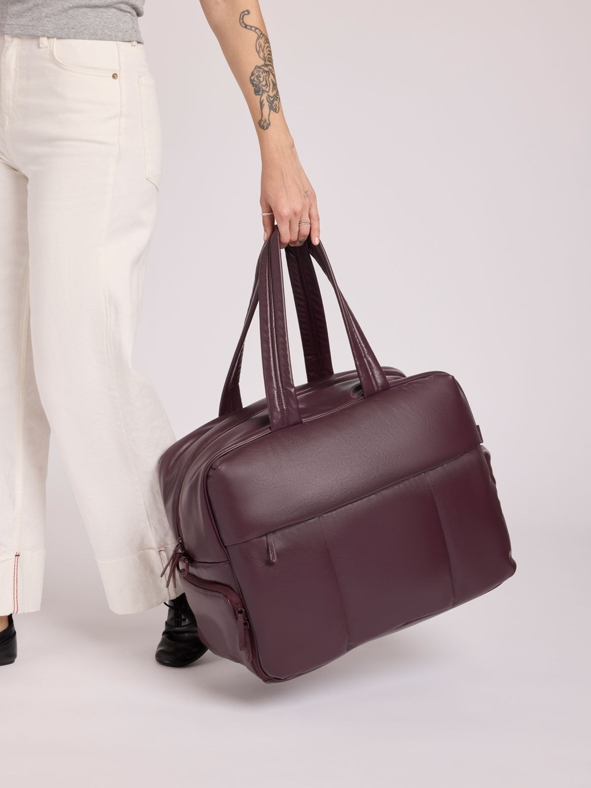 A person in white pants holds a large dark burgundy CALPAK Luka Duffel in vegan leather with dual top handles and a front zippered pocket, photographed against a clean white background; DLL2201-DARK-CHERRY-PU