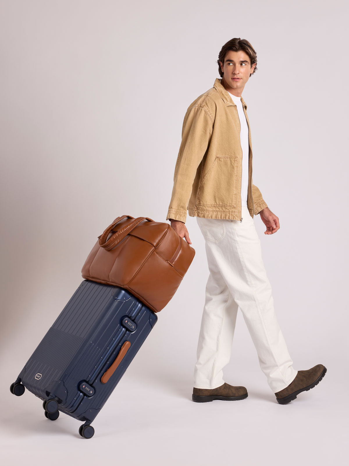 A man in a beige jacket, white shirt, and white trousers walks while pulling a CALPAK Vault Luggage suitcase with a Honey Brown CALPAK Luka Duffel in vegan leather with dual top handles and a front zippered pocket, photographed against a clean white background; DLL2201-HONEY-BROWN-PU