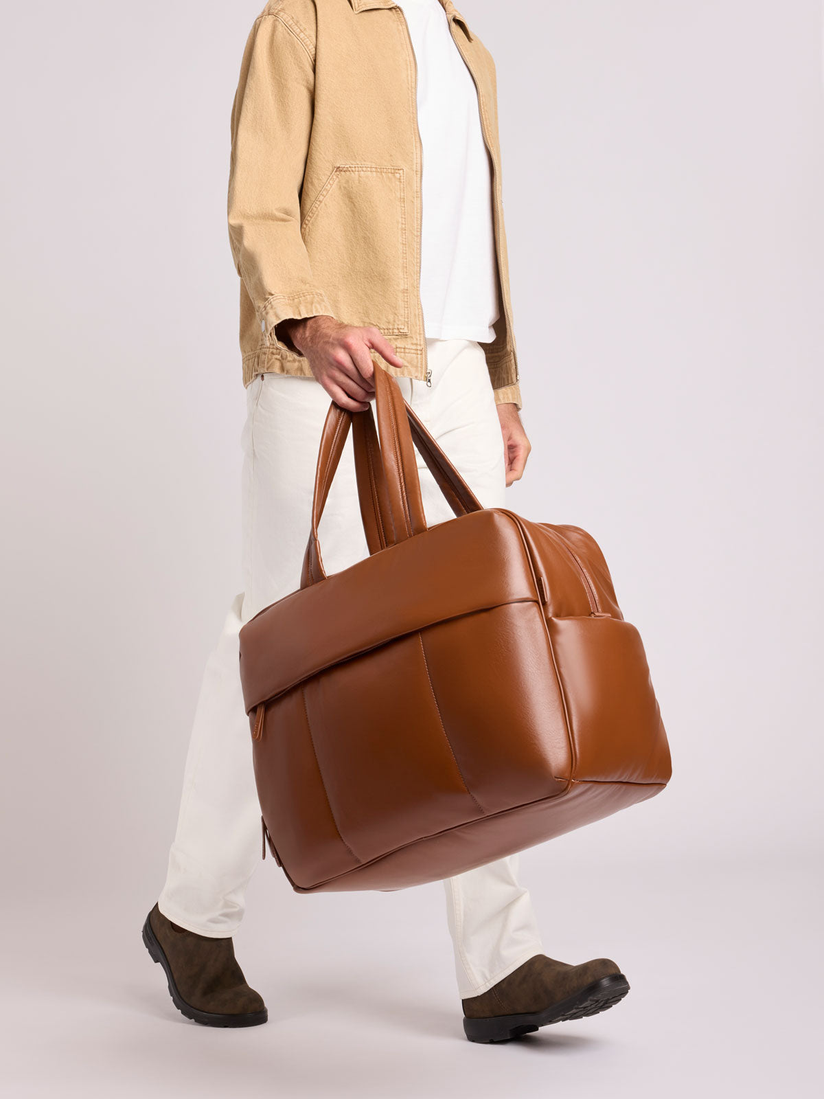A person wearing a beige jacket, white shirt, white pants, and dark brown shoes carries a Honey Brown CALPAK Luka Duffel in vegan leather with dual top handles and a front zippered pocket, photographed against a clean white background; DLL2201-HONEY-BROWN-PU