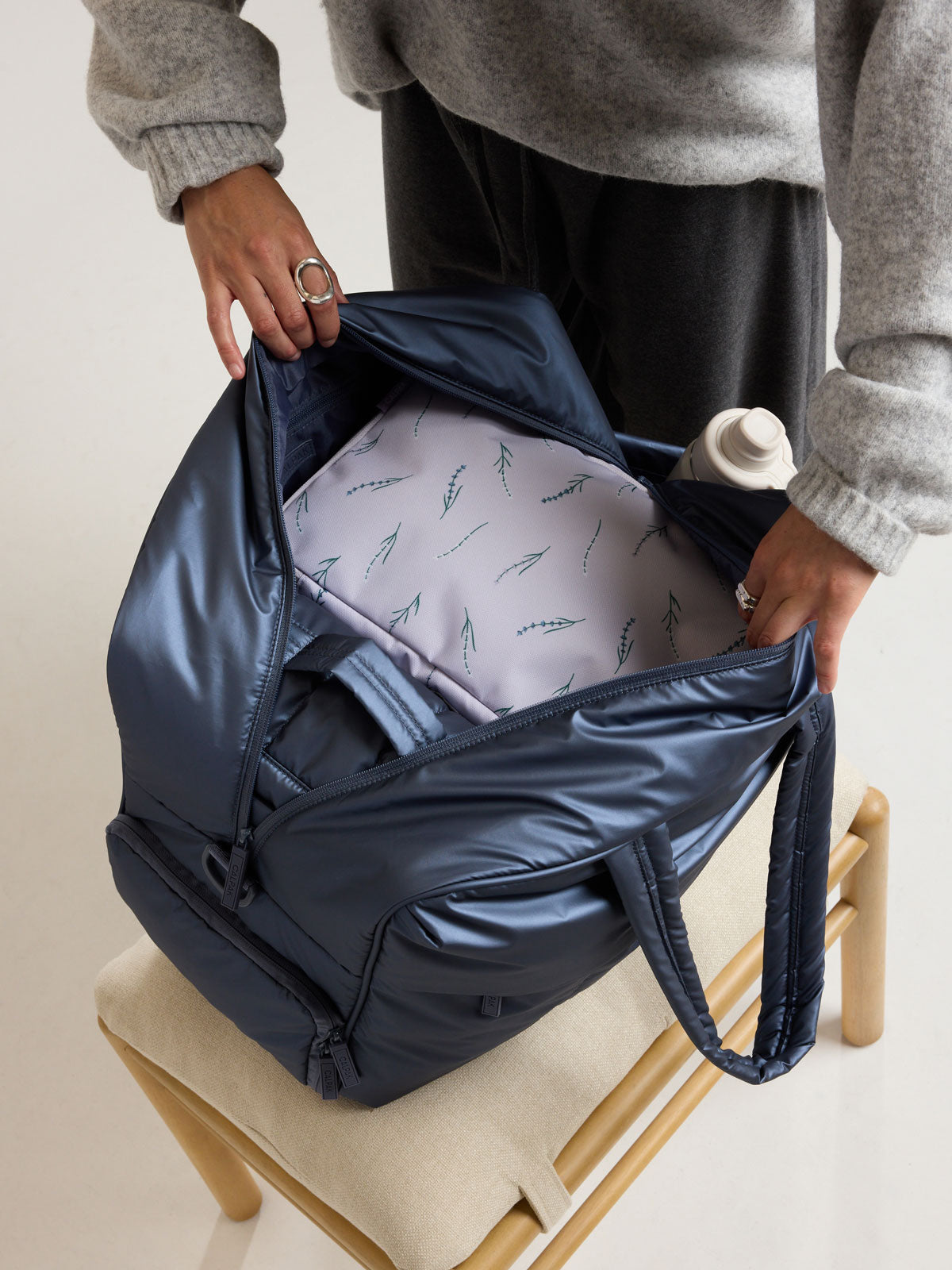 Model opening the CALPAK Luka Large Duffel in dark blue metallic material in Metallic Indigo; DLL2201-METALLIC-INDIGO