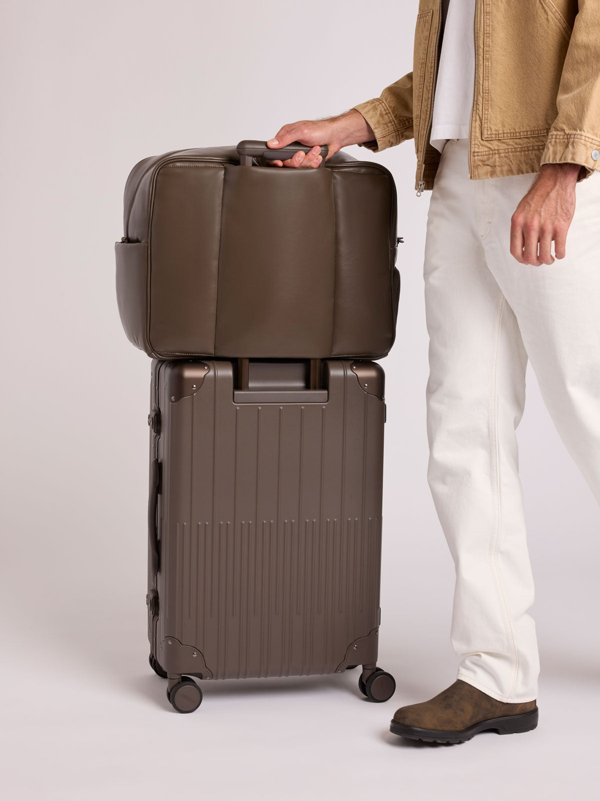 A person wearing a tan jacket, white pants, and brown shoes stands beside a dark brown CALPAK Vault Luggage with a matching dark brown Mushroom CALPAK Luka Duffel in vegan leather with dual top handles and a front zippered pocket, photographed against a clean white background; DLL2201-MUSHROOM-PU