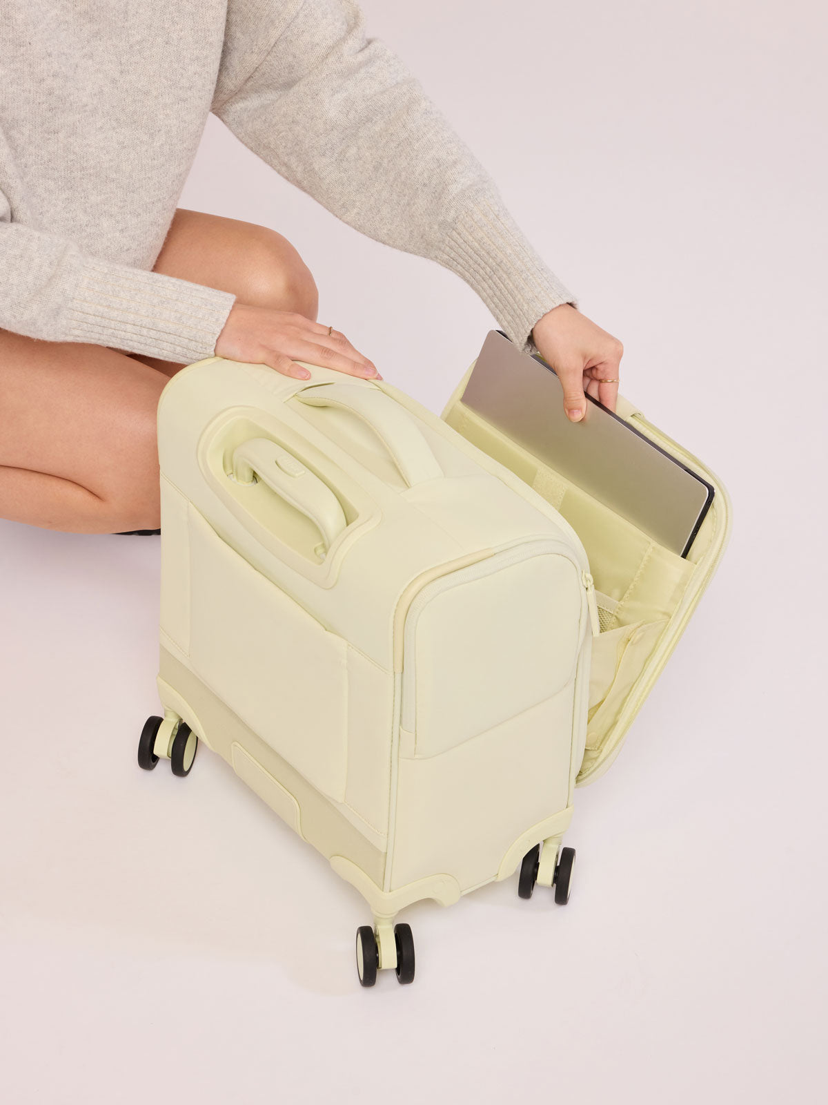 Model putting laptop in the laptop sleeve of CALPAK Luka Soft-Sided Mini Carry On Luggage in light green Celadon with spinner wheels and handle; LSM1014-CELADON