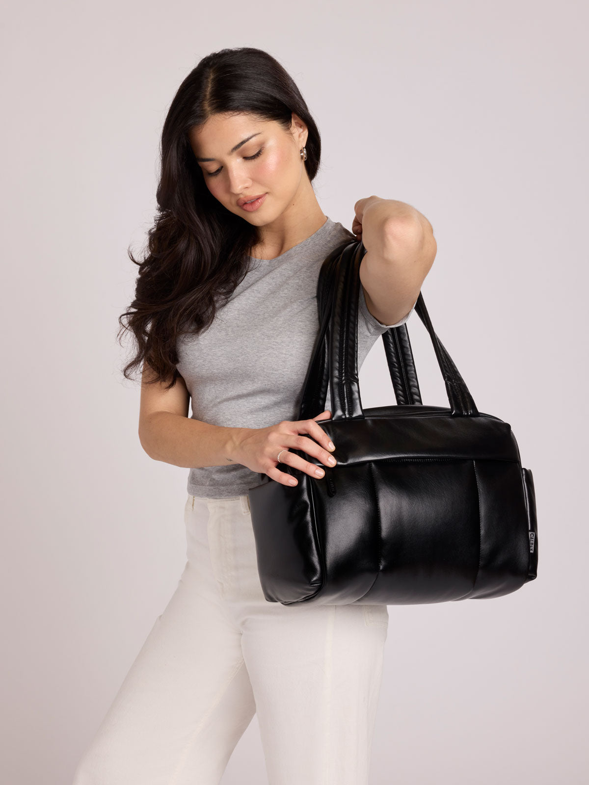 A woman wearing a gray t-shirt and white pants carries a black vegan leather CALPAK Luka Mini Duffel with dual top handles and side pockets, photographed against a clean white background; DLM2501-BLACK-PU