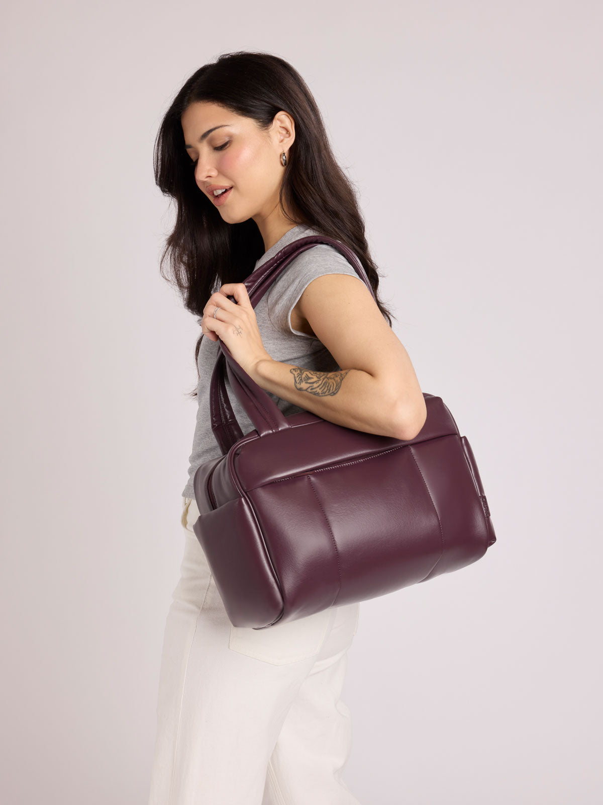 A woman with long dark hair is shown from the side, carrying a dark burgundy Dark Cherry vegan leather CALPAK Luka Mini Duffel with dual top handles and side pockets, photographed against a clean white background; DLM2501-DARK-CHERRY-PU