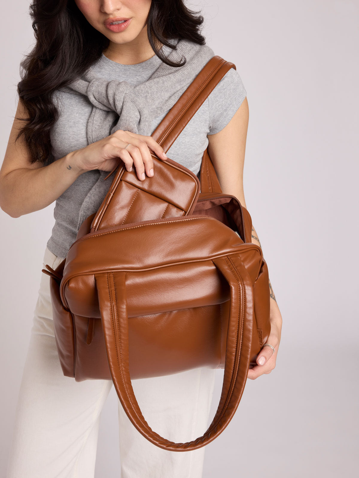 A woman wearing a gray t-shirt and white pants holds a Honey brown vegan leather CALPAK Luka Mini Duffel with dual top handles and side pockets, photographed against a clean white background; DLM2501-HONEY-BROWN-PU