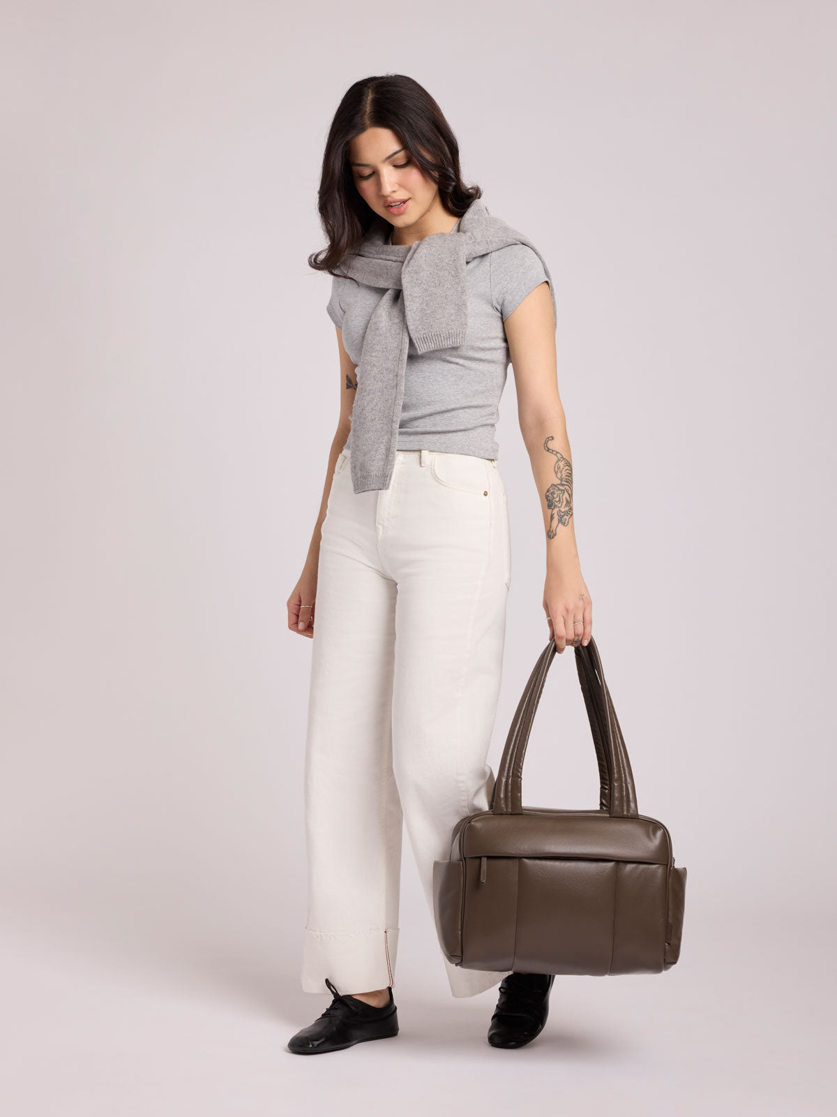 A woman modeling a light gray knit top with a large bow detail, white wide-leg pants, and black shoes, while holding a dark brown Mushroom vegan leather CALPAK Luka Mini Duffel with dual top handles and side pockets, photographed against a clean white background; DLM2501-MUSHROOM-PU
