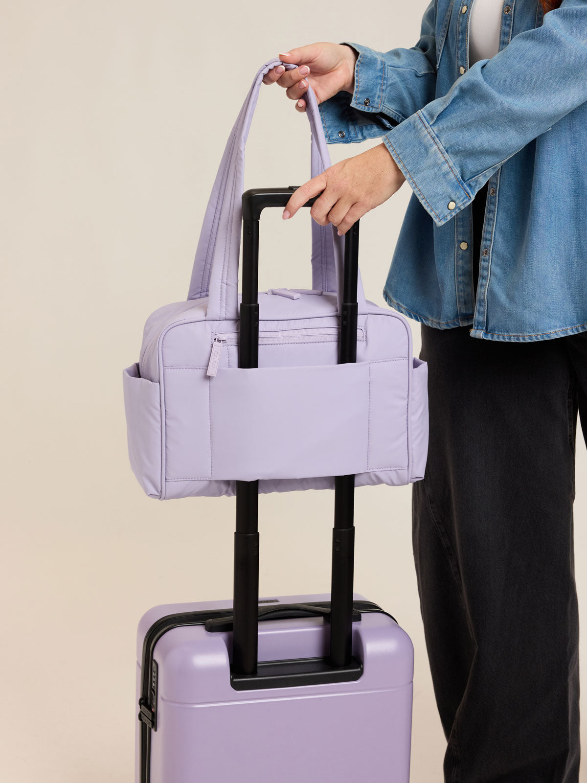 Model using trolley sleeve of CALPAK Luka Mini Duffel in light purple Orchid on a white background; DLM2501-ORCHID