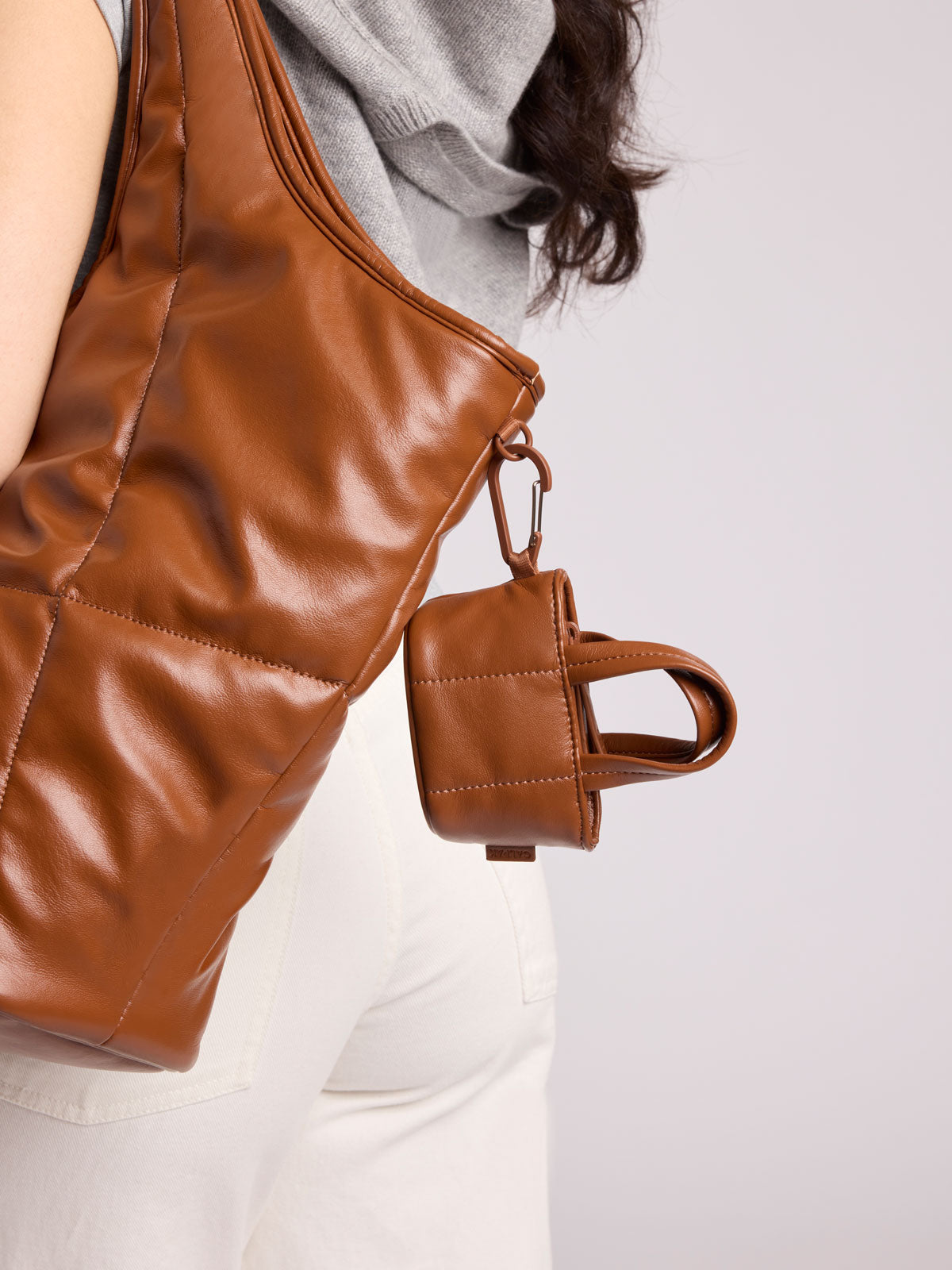A close-up of a person wearing a large CALPAK Everything Tote in Honey Brown Vegan Leather with a matching small CALPAK Luka Tote Zip Bag Charm attached by a metal ring; AMT2601-HONEY-BROWN-PU