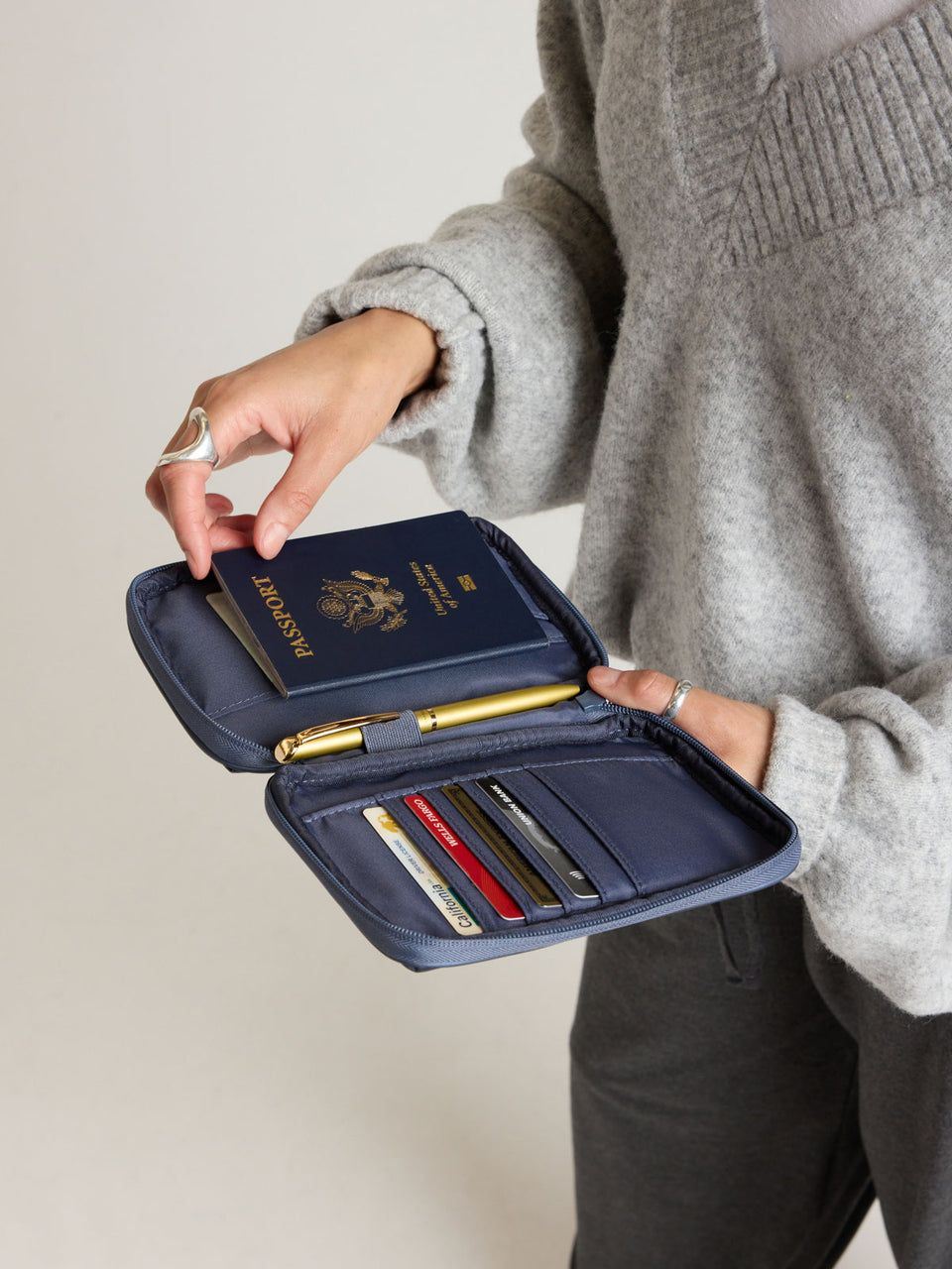 Model putting passport into the CALPAK Luka Zippered Passport Wallet in dark blue metallic material in Metallic Indigo; PZW2301-METALLIC-INDIGO