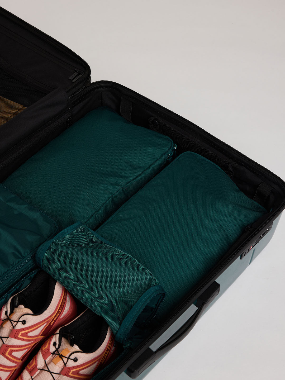 Two dark green teal Kale CALPAK Medium Compression Packing Cubes arranged inside an open black suitcase, with a pair of pink and black sneakers visible in the lower left compartment; PCC2201-KALE