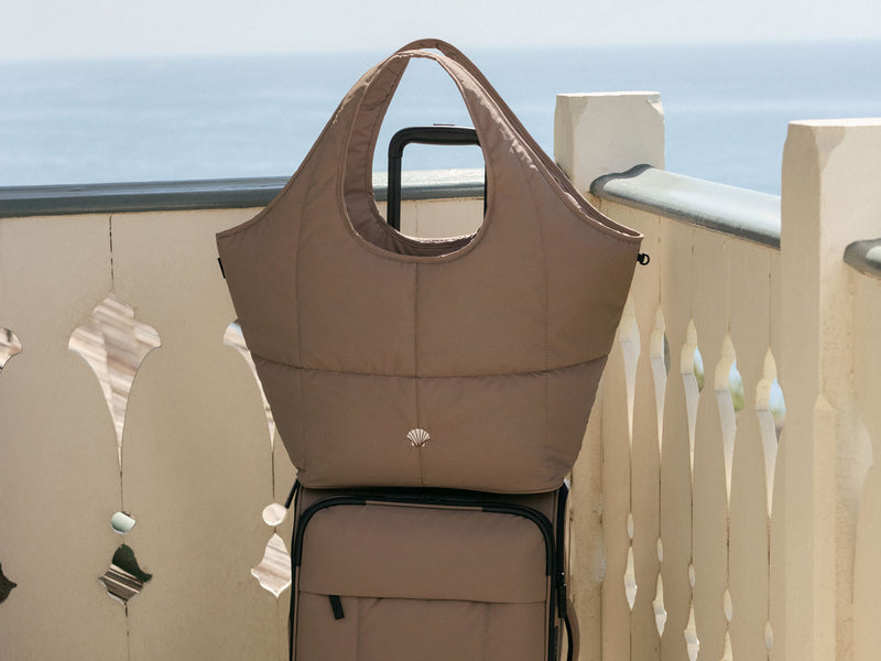 Brown CALPAK Luka Soft Sided Carry On Luggage in Chocolate with a matching brown Chocolate Luka Everything Tote bag on a balcony with ocean view