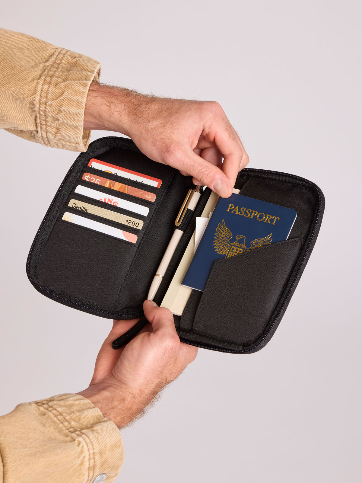 A CALPAK Luka Passport Wallet in Black Vegan Leather shown open, held by a person's hands. Inside, card slots hold credit cards, a pen, and a blue passport is tucked into the side pocket; PZW2301-BLACK-PU