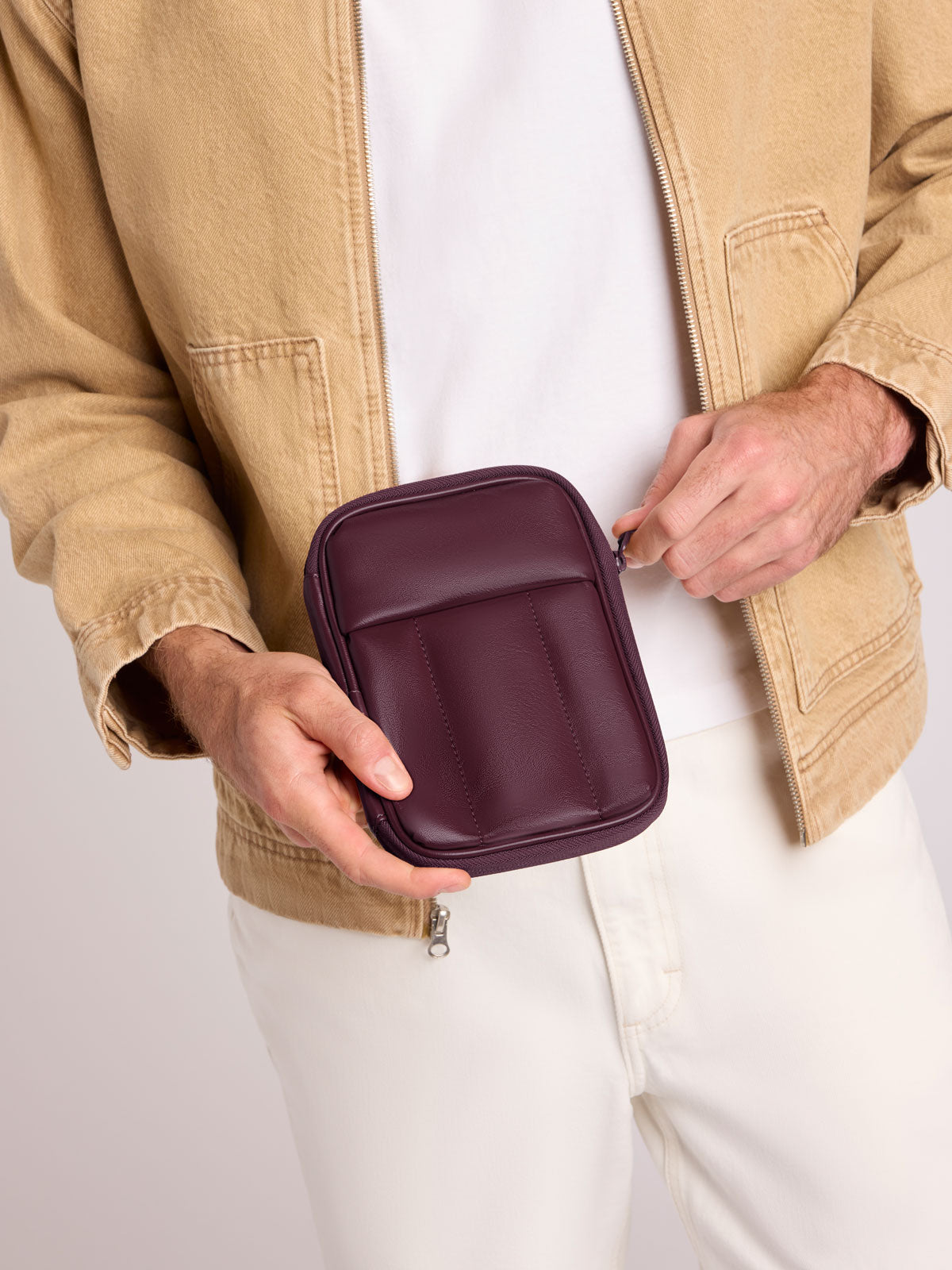 A person wearing a tan jacket and white pants holds a CALPAK Luka Zip Passport Wallet in Dark Cherry Vegan Leather against a plain light background; PZW2301-DARK-CHERRY-PU