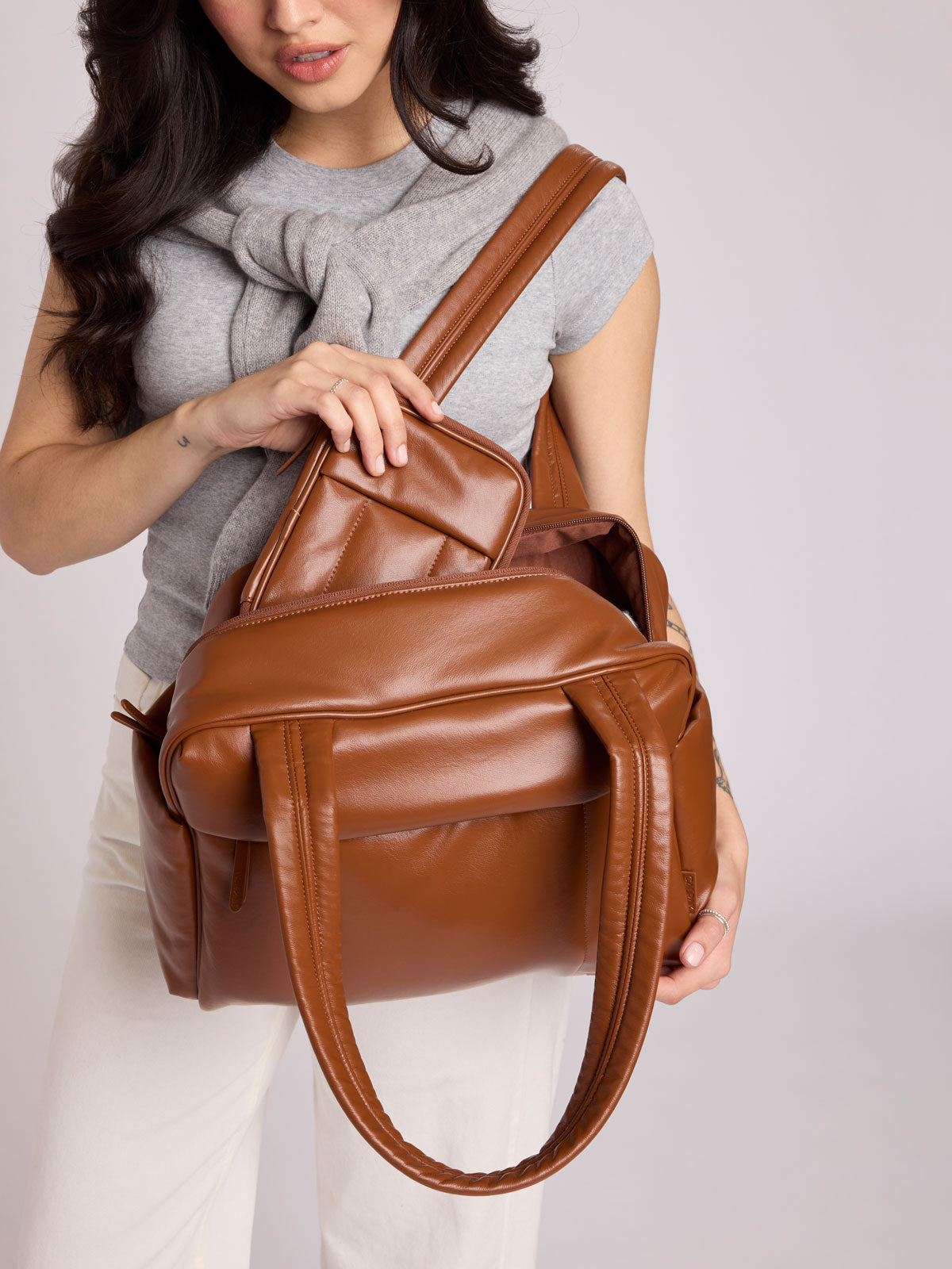 A woman wearing a gray knit top and white pants holds a CALPAK Luka ZIp Passport Wallet in Honey Brown Vegan Leather with matching Luka Mini Duffel against a light gray studio background ;PZW2301-HONEY-BROWN-PU