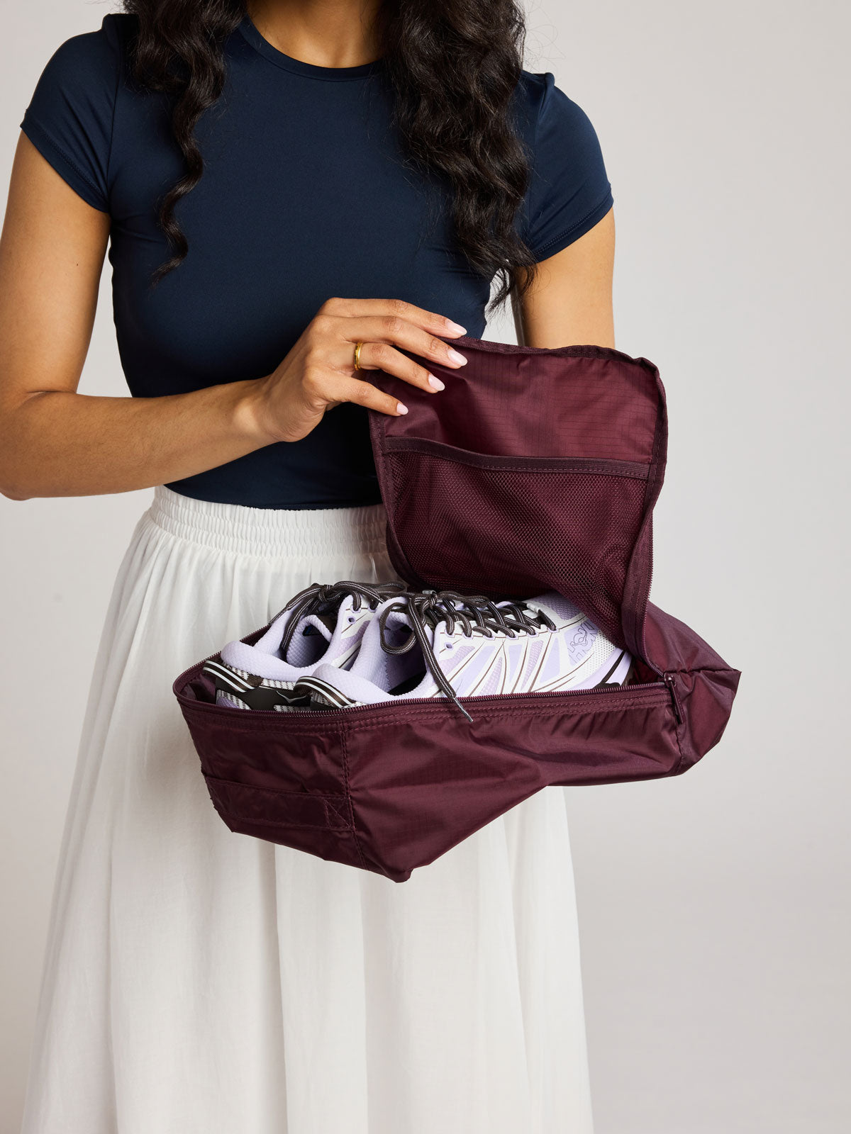 A woman wearing a navy blue top and white skirt holds an burgundy Dark Cherry rectangular CALPAK shoe cube with zippers and top handle shown on a plain background; KSB2001-DARK-CHERRY