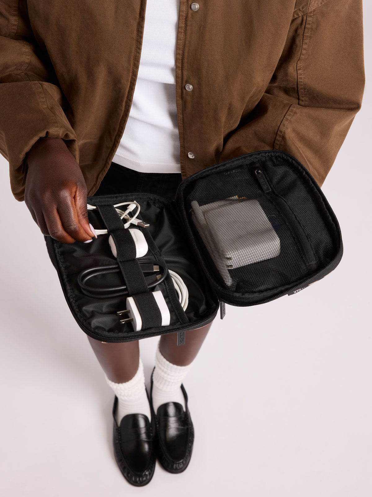 A person wearing a brown jacket and white shirt holds open CALPAK Tech Organizer pouch in Leopard containing a power strip, cables, and a gray power bank, against a light gray studio background;  ATC2001-LEOPARD