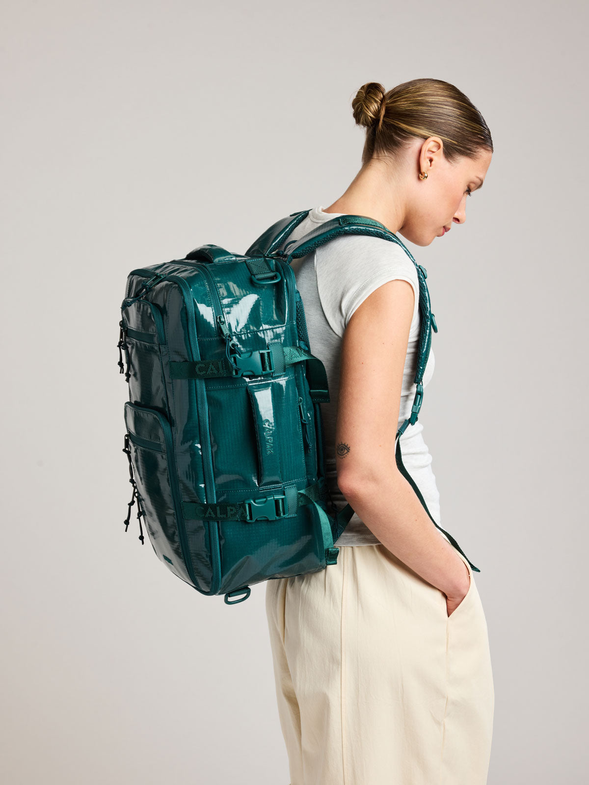 Model in a white shirt wearing the CALPAK Terra 26L Laptop Backpack in dark blue green Kale with multiple pockets on a white background; BPH2201-KALE