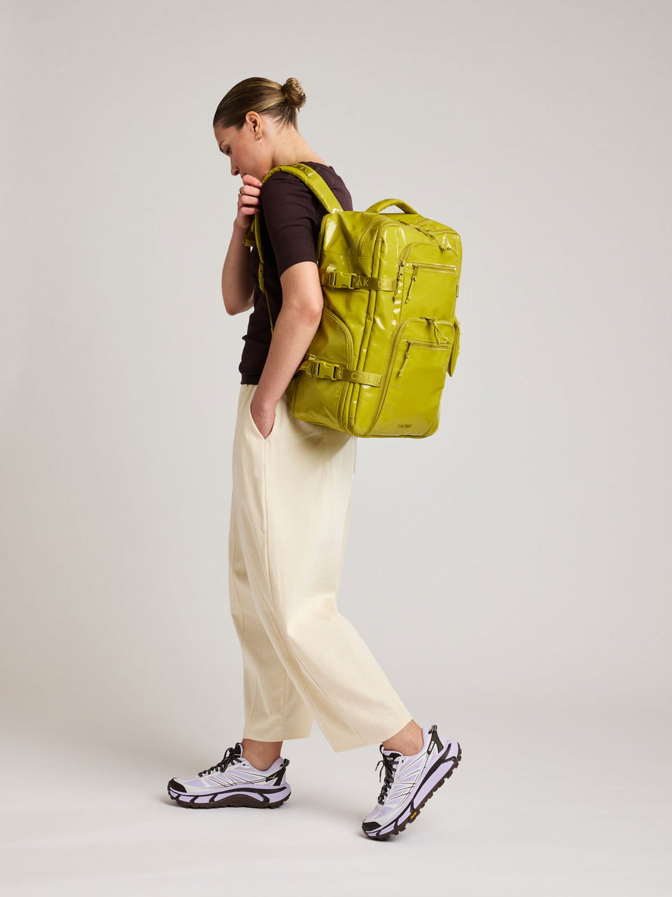 Person with a brown top wearing the CALPAK Terra 26L Laptop Backpack in yellow green Wasabi with multiple pockets on a white background; BPH2201-WASABI