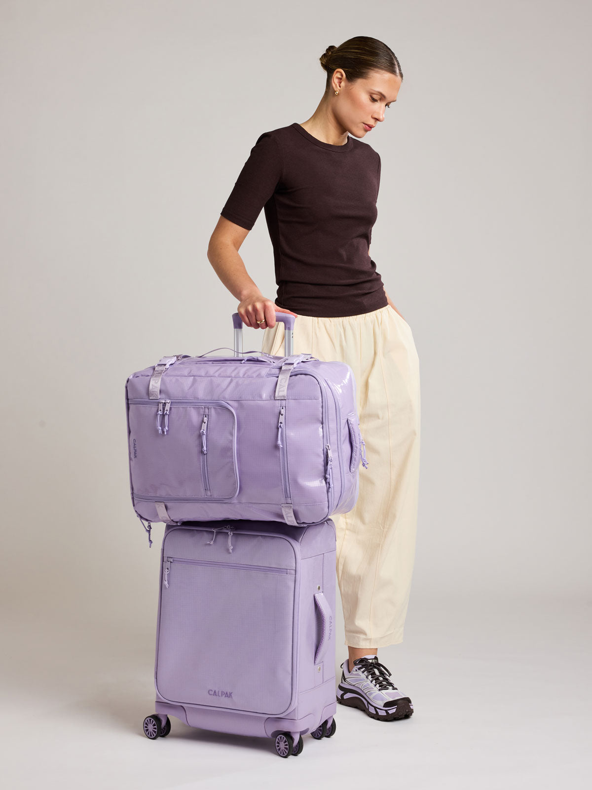 Woman with suitcase and the CALPAK Terra 35L Laptop Backpack Duffel in lavender Amethyst on top of suitcase with a white background; BPTL2601-AMETHYST