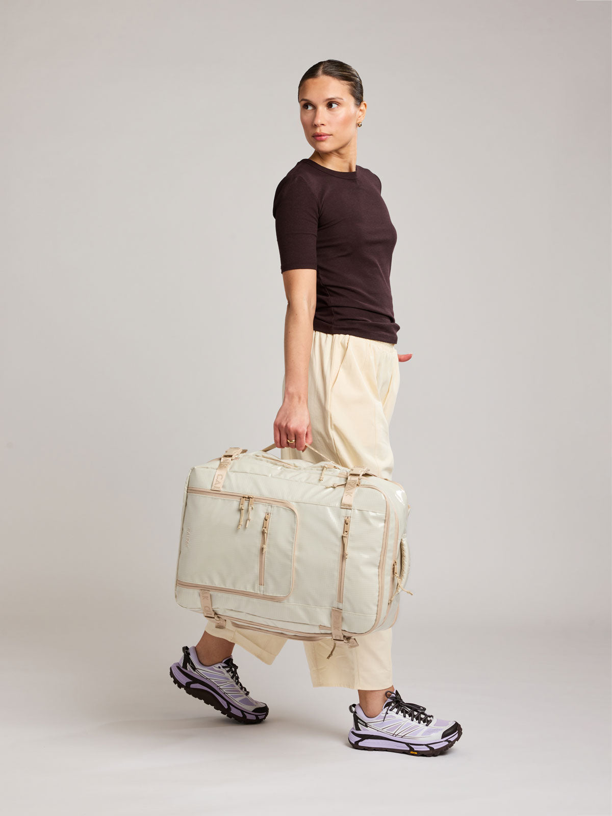Woman holding the side handle of the CALPAK Terra 35L Laptop Backpack Duffel in off-white White Sands with tan accents on a white background; BPTL2601-WHITE-SANDS