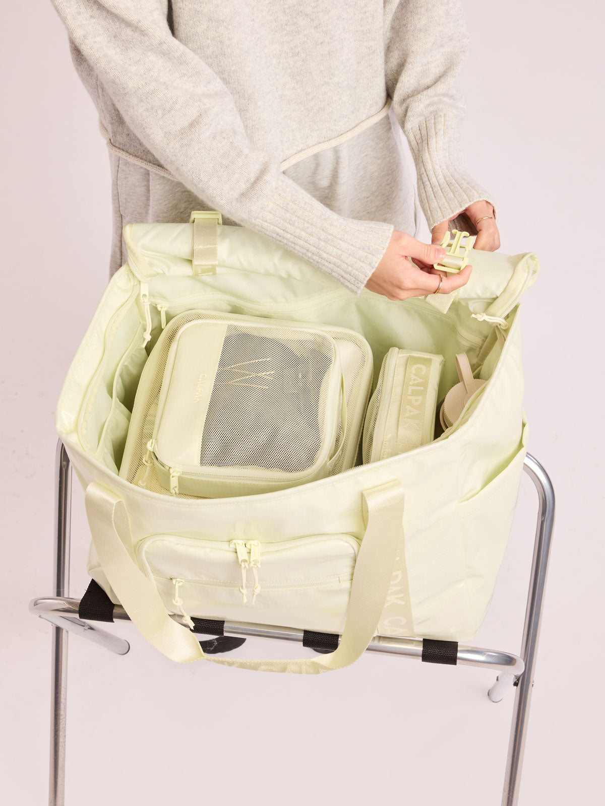 Model adjusting straps of the CALPAK Terra 35L Tote Bag with multiple pockets and shoulder straps in shiny light green Celadon on a white background; TTO2401-CELADON