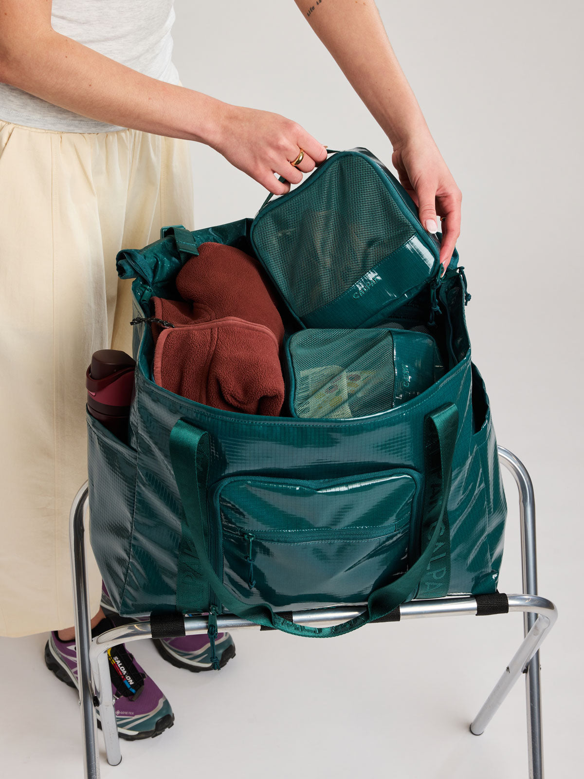 A dark teal green Kale travel  CALPAK Terra 35L Tote bag with a front zippered pocket, being packed with toiletry bags by a person; TTO2401-KALE