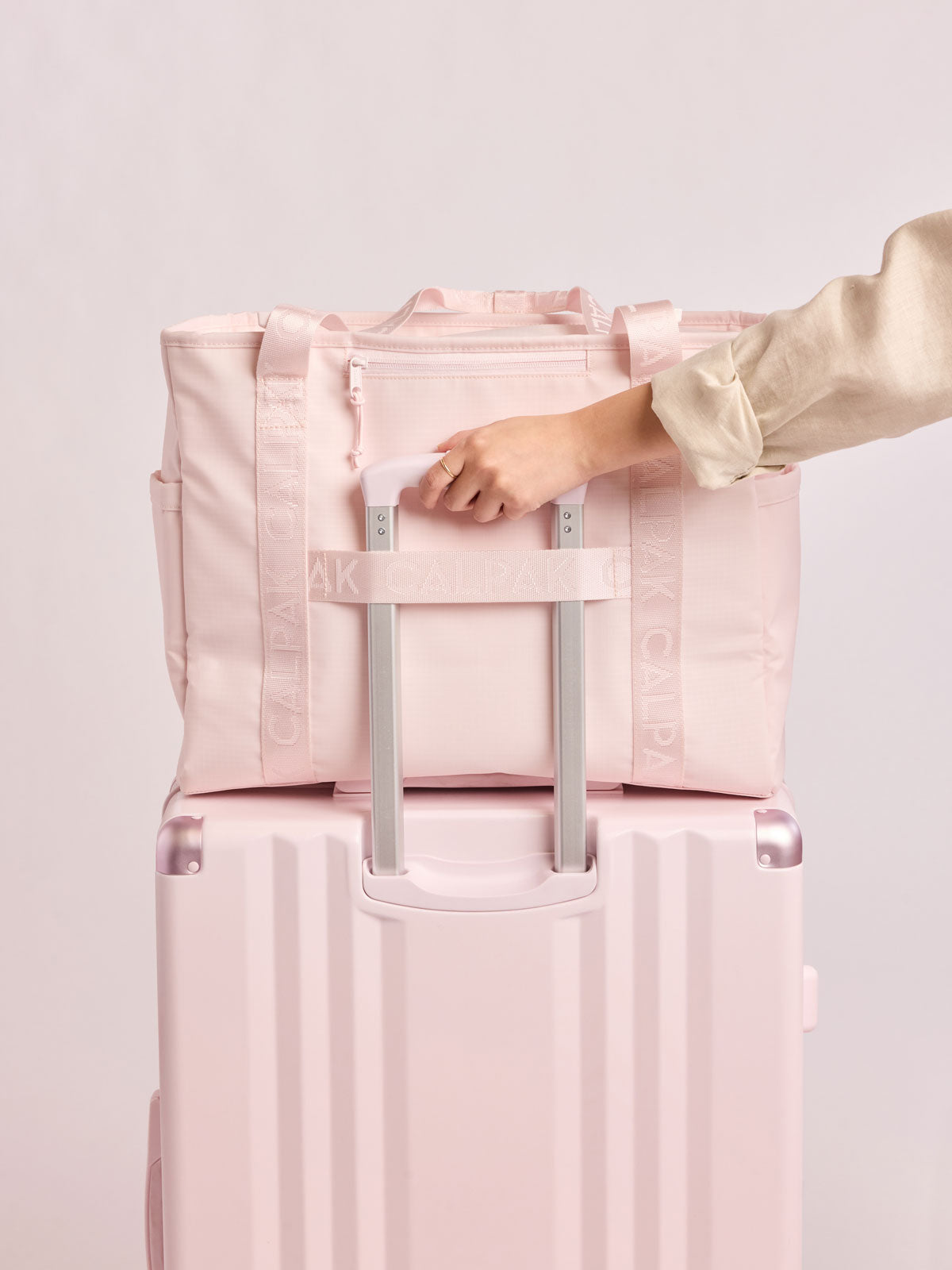 Model holding a luggage with the CALPAK Terra 35L Tote Bag with multiple pockets and shoulder straps in shiny light pink Shell Pink on a white background and luggage trolley sleeve; TTO2401-SHELL-PINK