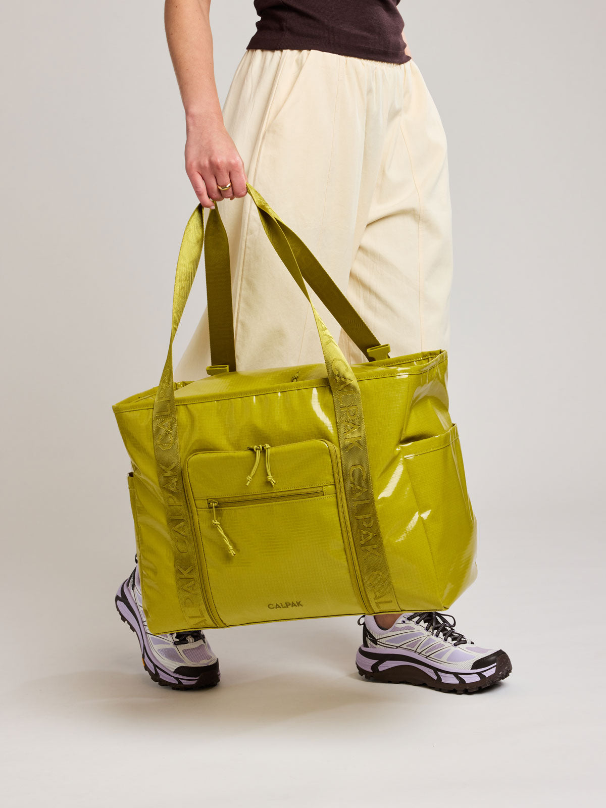 A person wearing cream-colored pants and athletic shoes holds a large, shiny lime green Wasabi CALPAK Terra 35L Laptop tote bag with dual shoulder straps against a plain light gray studio background; TTO2401-WASABI