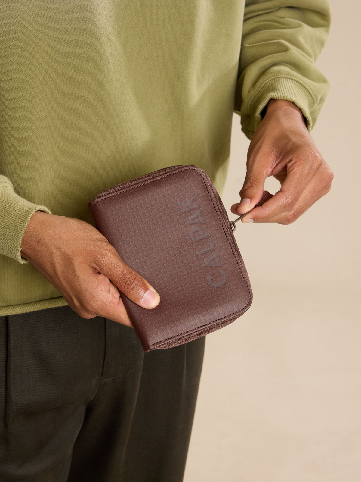 Model zipping up the CALPAK Terra Zippered Passport Wallet in brown Walnut front view; ATPW2501-WALNUT