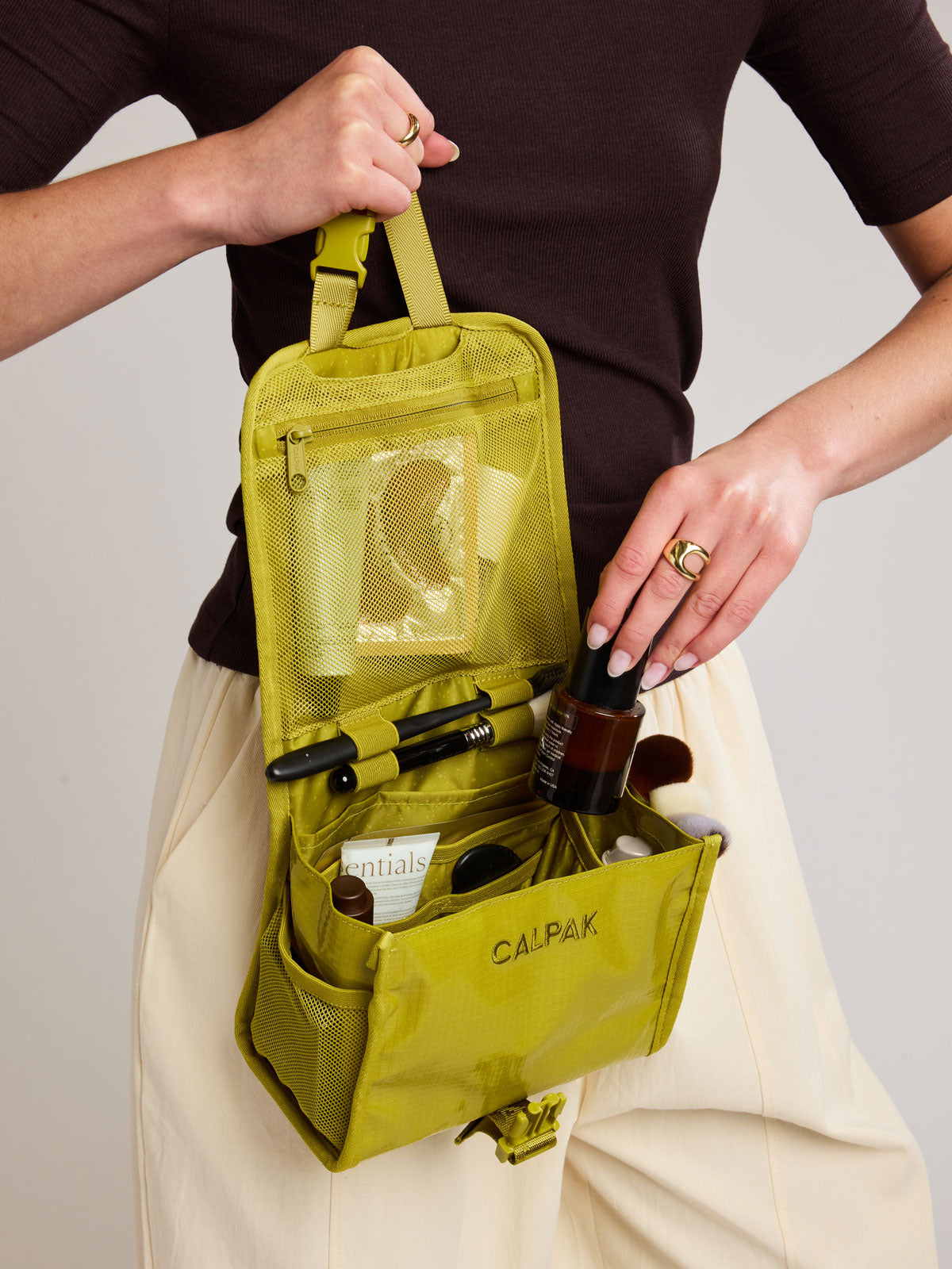 Model adding toiletry into CALPAK Terra Hanging Toiletry Bag to store makeup and toiletries in yellow green Wasabi on a white background; THY2201-WASABI