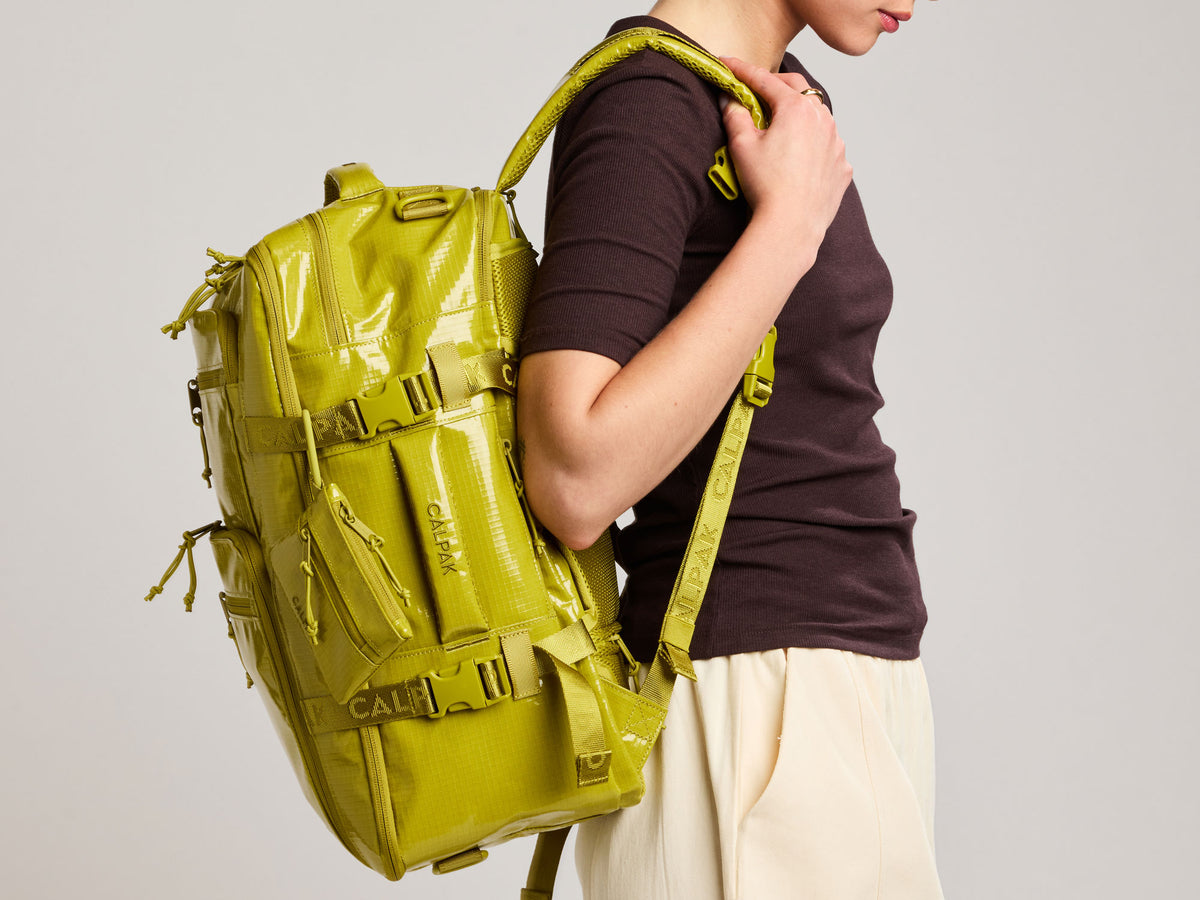 A person wearing a dark brown t-shirt and cream pants carries a lime green Terra 35L  backpack with multiple straps and buckles against a light gray background.
