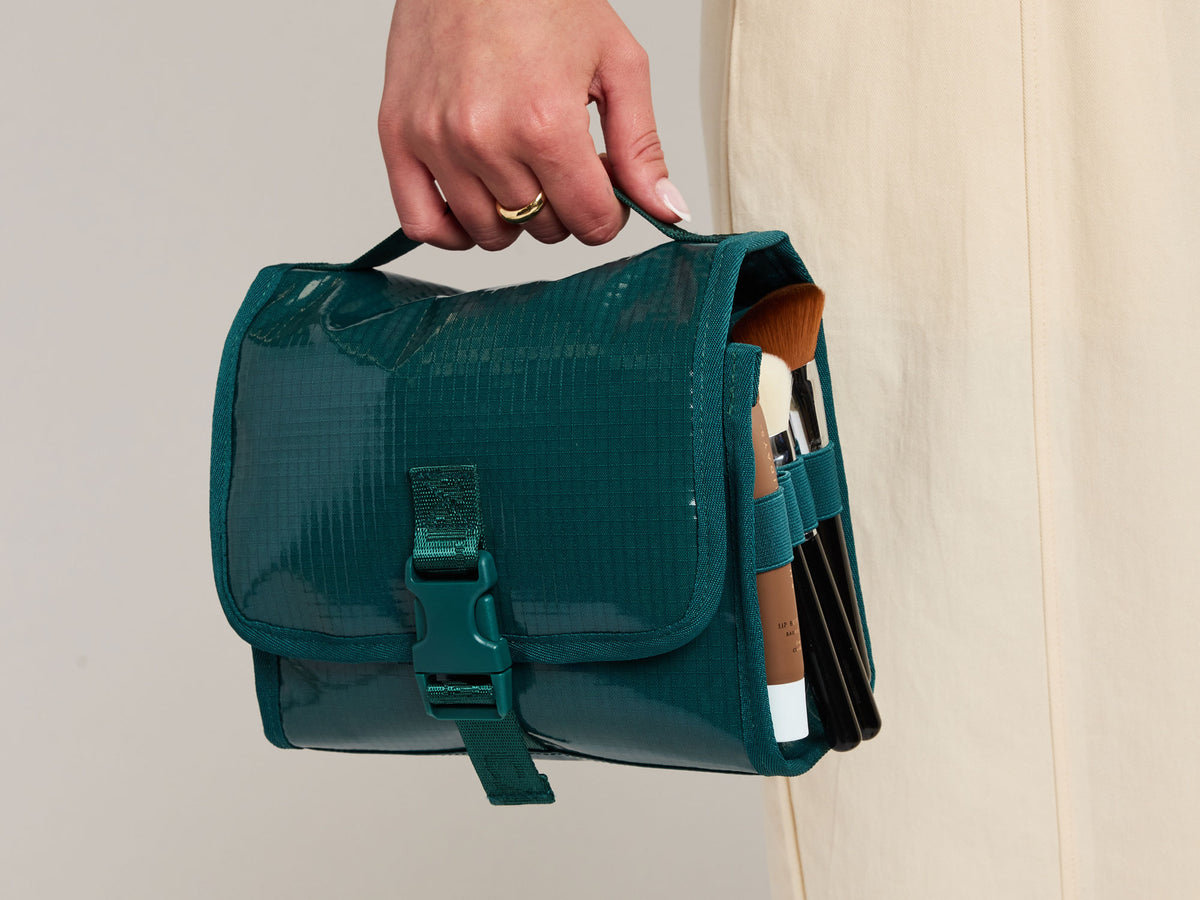 A hand holding a dark teal green Terra waterproof makeup bag with a buckle closure and side pockets containing makeup items, set against a neutral beige background.